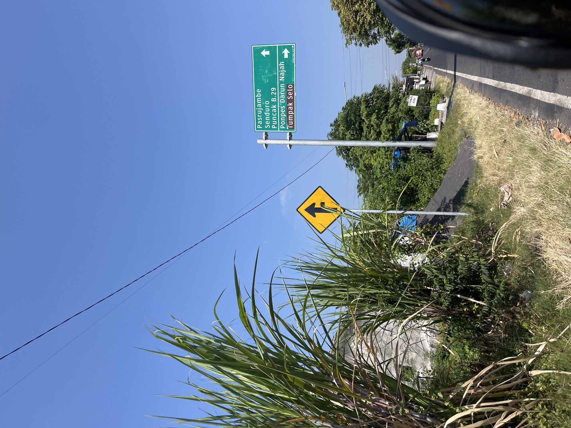 The image captures a roadside scene in Purworejo, Indonesia, under a clear blue sky. In the foreground, lush green sugarcane stalks dominate the left side of the frame, with dry grass and some debris visible on the roadside verge. A yellow diamond-shaped traffic sign with a black arrow pointing upwards is positioned on a pole.
Further down the road, a green directional signpost stands. It displays destinations in white text: "Pasrujambe Senduro", "Puncak B.29", "Ponpes Darun Najah", and "Tumpak Selo". Arrows indicate the directions for these locations. To the right of the signpost, the road stretches into the distance, with a few vehicles, including motorcycles and a truck, visible. Trees and vegetation line the sides of the road, with a few small structures and a blue tarp also present. The perspective suggests the photo was taken from a moving vehicle, with a portion of a black helmet visible in the bottom right corner.