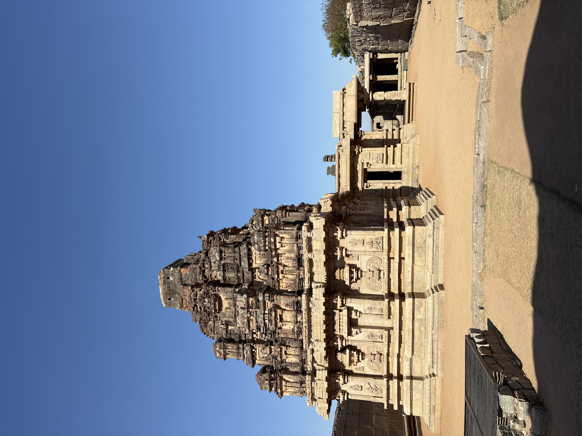 The image shows the Hazara Rama Temple in Kamalapura, India. The structure is a large, ornate stone building with multiple tiers and detailed carvings. The sky is clear and blue. The surrounding area appears to be dry and dusty, with other stone structures in the background.