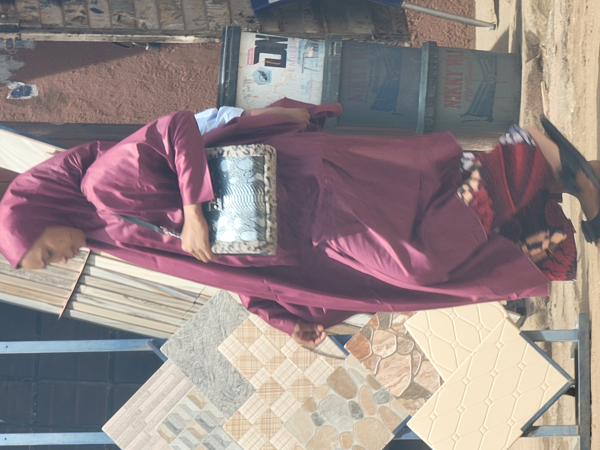 A woman in a maroon headscarf and gown is standing in front of a display of tile samples. She is holding a purse or handbag with a silver or gray sequined design. The tiles have various textures, colors, and patterns. Behind the woman and tiles, there are stacks of what appear to be sealed boxes or containers. The wall behind these containers is red-brown. The location appears to be outdoors, perhaps a tile store or showroom in Maiburiji, Nigeria.