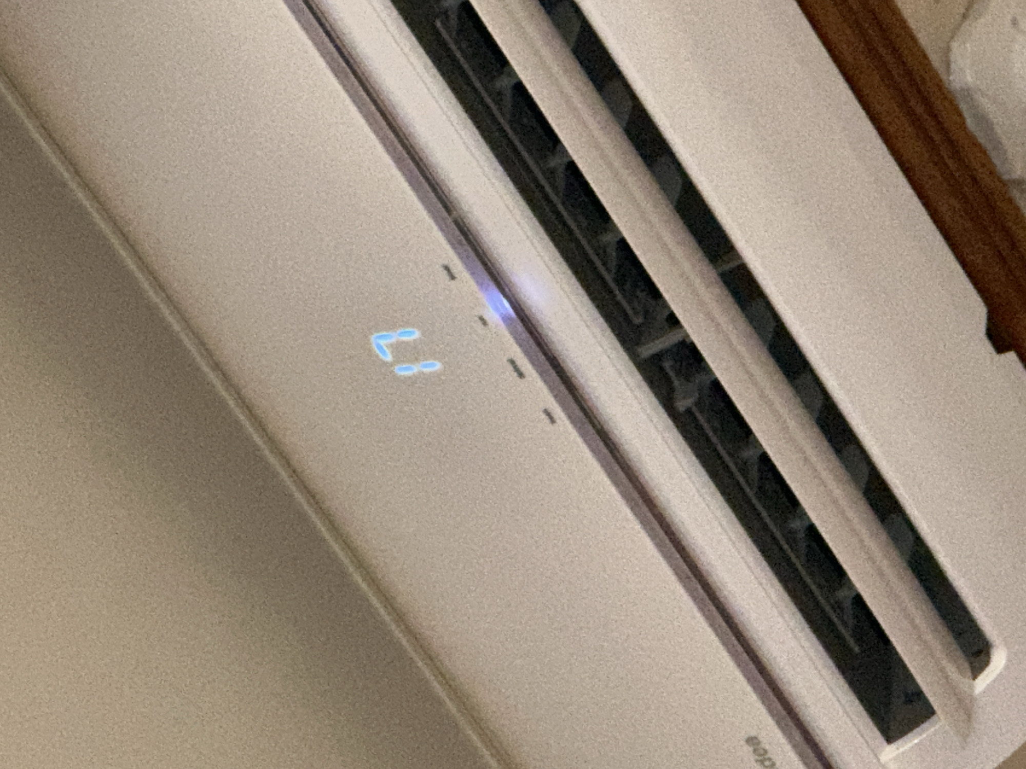The image shows a close-up view of an air conditioner unit, likely indoors, displaying a temperature of 17 degrees. The unit is white and has a digital display showing the number "17" in blue LED lights. There are also three small indicator lights visible above the display. The louvers of the air conditioner are visible, suggesting it is either on or has recently been used. The background appears to be a textured wall. The brand name "Midea" is partially visible on the bottom left of the unit. The image is taken from a slightly upward angle, focusing on the front panel of the air conditioner. The overall lighting is dim, with the blue display being the brightest element.