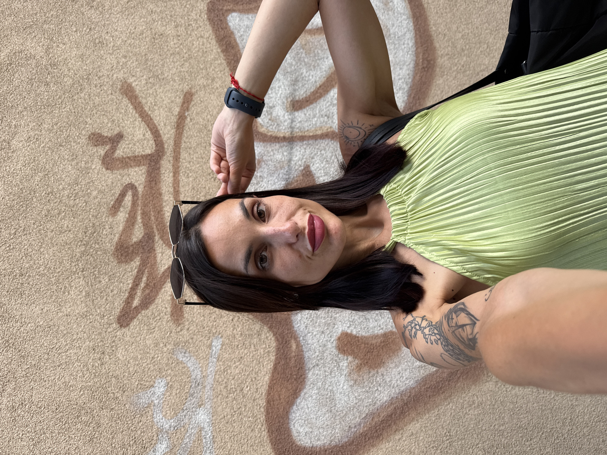 A woman is posing for a selfie in front of a graffiti-covered wall. She has dark hair, sunglasses perched on her head, and is wearing a light green pleated halter top. She is also wearing a dark watch on her left wrist with a red string tied around it. Tattoos are visible on her left arm. The wall behind her is textured and appears to be made of concrete, with brown and white graffiti spray-painted onto it. The setting appears to be an outdoor urban environment.