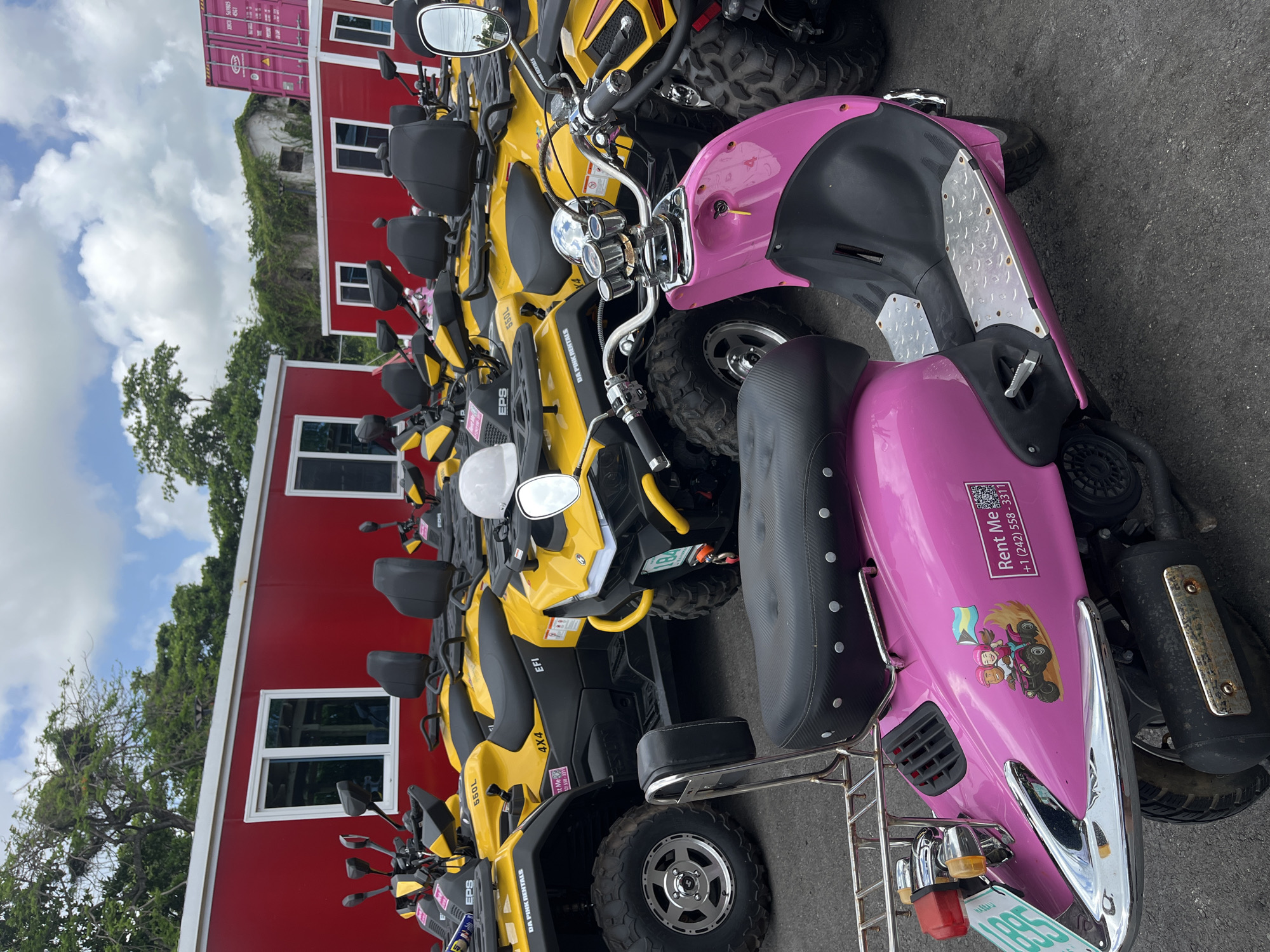The image shows a row of yellow ATVs parked next to a pink scooter. The ATVs are branded "DA PINK RENTALS" and "EPS 550L". The scooter has a sticker with a QR code and a phone number "+1 (242) 558-3311", along with the words "Rent Me". Behind the vehicles is a red building with white-framed windows. In the background, there is a shipping container and a building covered in vines. The location is Unknown, Unknown.
