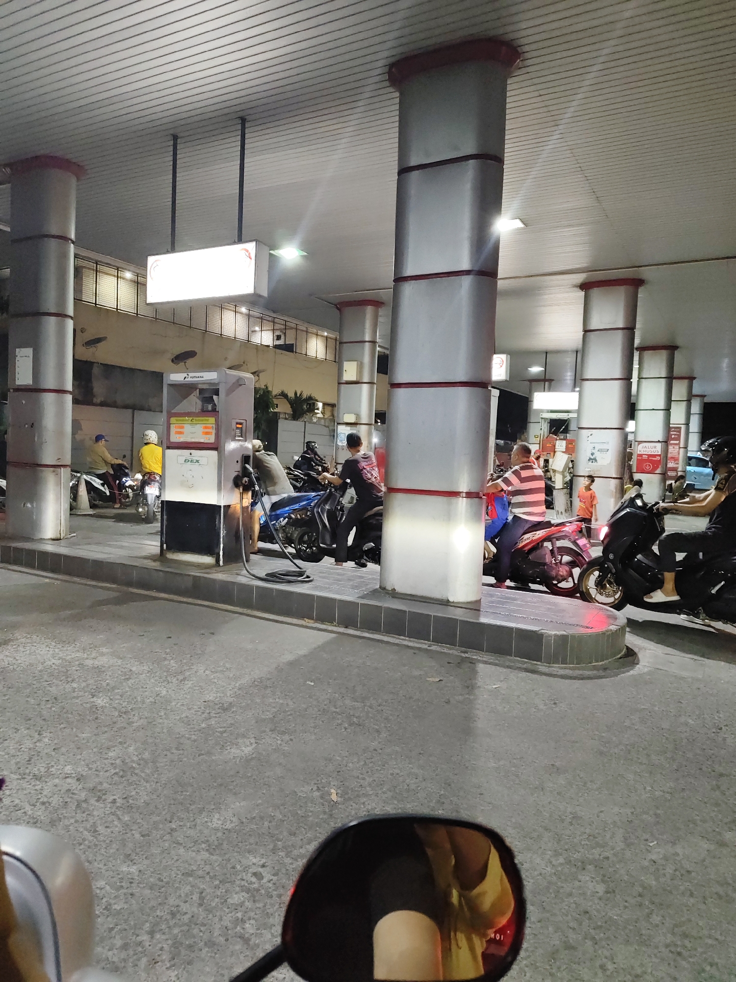 The image depicts a nighttime scene at a Pertamina gas station in South Jakarta, Indonesia. The gas station features a covered area with multiple large, silver pillars supporting a corrugated metal roof. Several motorcycles are present, with riders refueling or waiting in line. A prominent gas pump with a digital display is visible, labeled "DEX". People are seen in various poses, some wearing helmets. The ground is paved asphalt. In the foreground, a motorcycle's rearview mirror reflects a person wearing a yellow shirt. The overall atmosphere suggests a typical, active gas station environment in the specified location.
