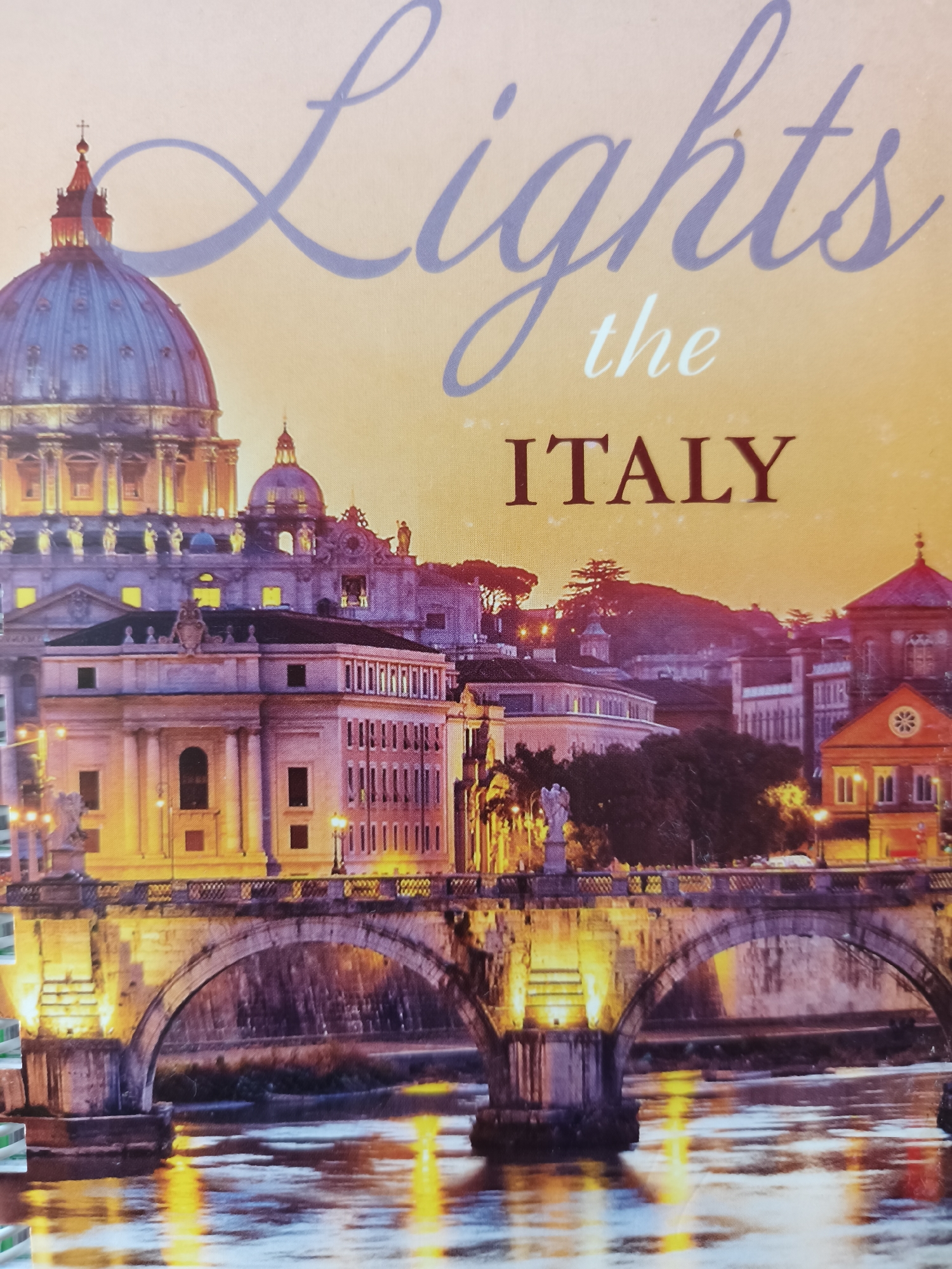 This media file captures a vibrant cityscape at dusk or night, featuring prominent architectural landmarks and a river. The central focus is a majestic dome, clearly identifiable as St. Peter's Basilica in Vatican City, Rome, Italy, glowing with blue and purple tones, while the surrounding structure is illuminated with warm yellow light.
Below the basilica, an ornate stone bridge with multiple arches spans a river, its features and railings brightly lit and casting shimmering golden reflections onto the water's surface. Numerous other buildings with classical architecture, including columns, balconies, and windows, line the riverbanks and recede into the background, also lit up to reveal their intricate details. The sky above displays a gradient of warm orange and yellow near the horizon, fading into deeper purples and blues, indicative of twilight or early night. The overall scene evokes a sense of grandeur and tranquility.
Visible text in the upper portion of the image reads: "Lights" in a large, elegant script, followed by "the" in smaller text, and finally "ITALY" in bold, capitalized letters. The depicted location is unequivocally Rome, Italy, despite the provided location context of Kryvyi Rih, Ukraine. No specific activity or people are discernible; the image focuses on the illuminated urban landscape.
