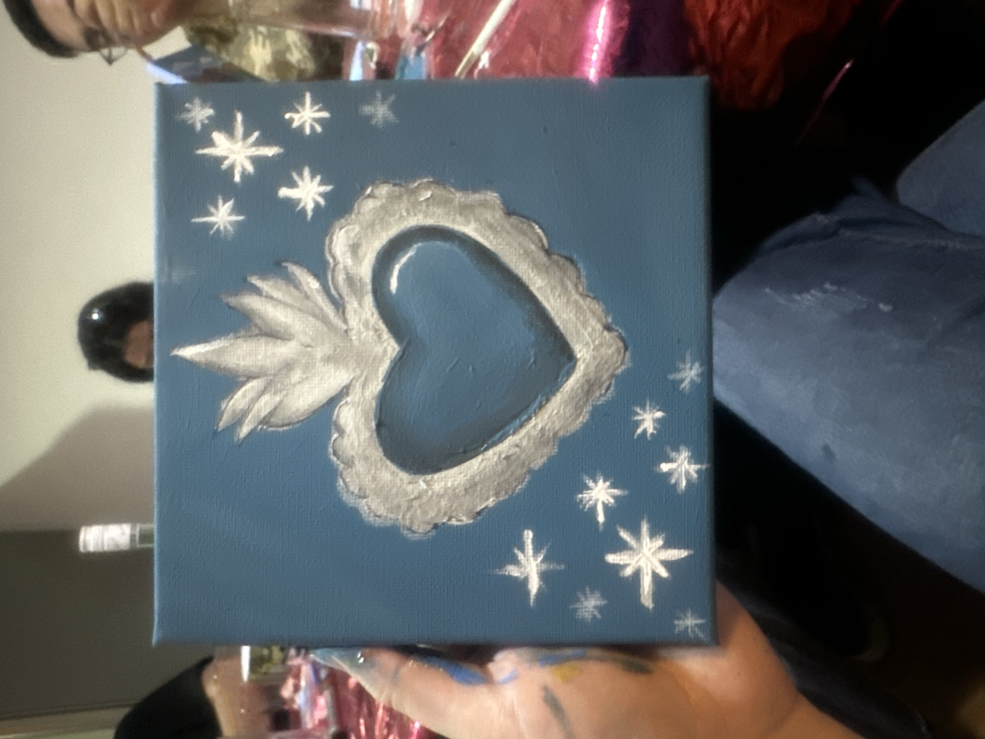 A person is holding a small, square, blue canvas painting. The painting depicts a heart shape with a stylized, radiating design and several star-like shapes surrounding it.