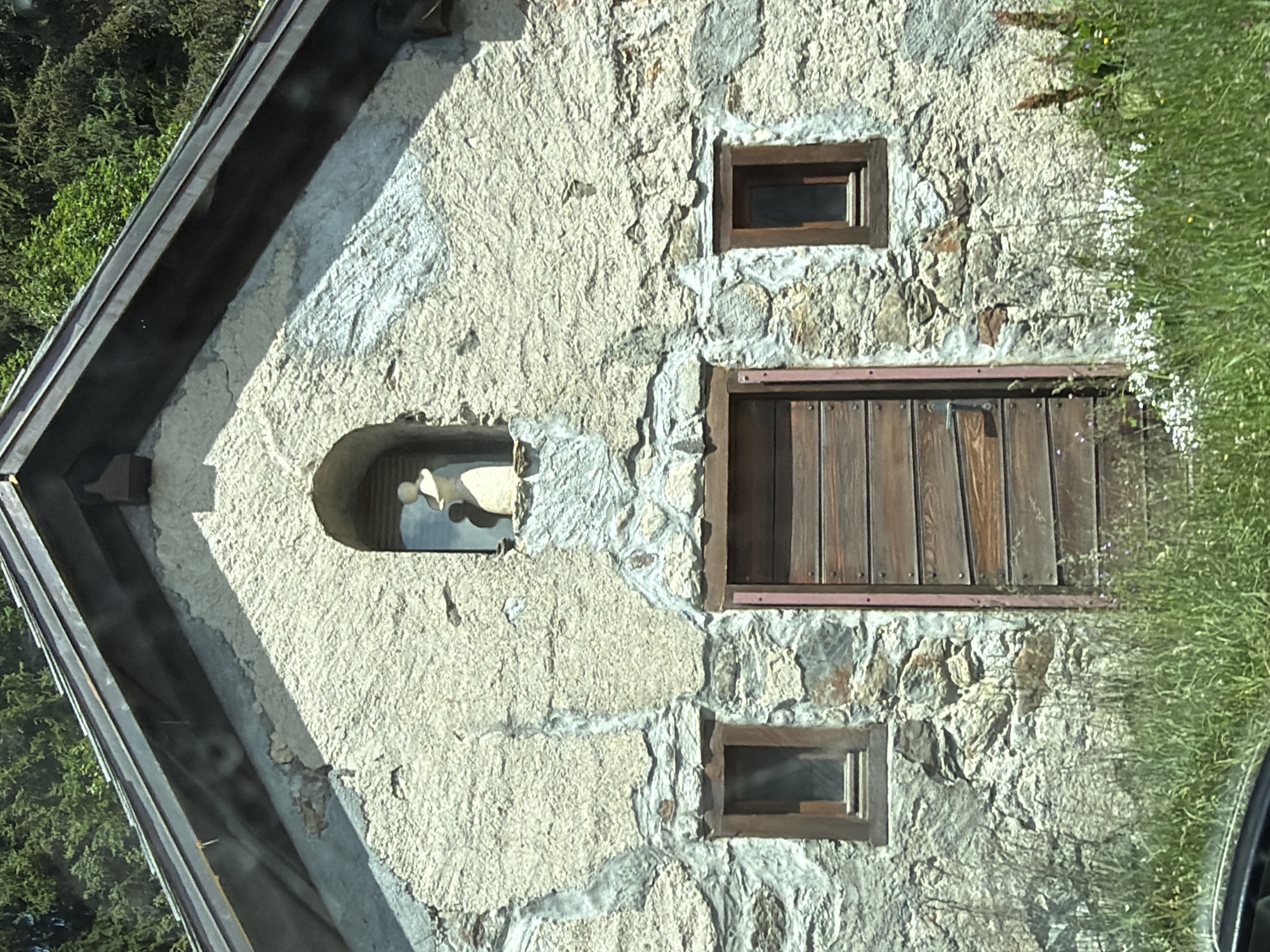The image shows the front facade of a stone and stucco building, likely a small chapel or hermitage, set against a backdrop of green trees. The building has a pitched roof covered with dark material. A small, arched niche is centered on the upper part of the facade, containing a light-colored statue of a person wearing a hat. Below the niche, there is a large wooden door with horizontal planks. Two small windows with wooden frames are located on either side of the door, one on the left and one on the right. The walls are constructed of rough-hewn stones and a textured stucco finish. The foreground is overgrown with tall green grass and scattered wildflowers. The scene appears to be located in a rural or mountainous area, consistent with the setting of Vallorcine, France.