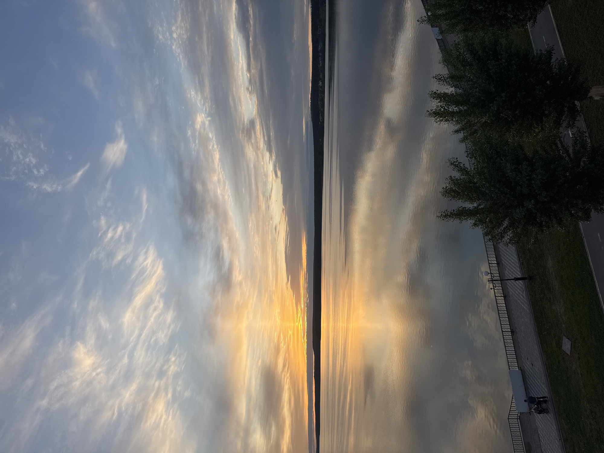 The image shows a sunset over the Votkinsk Reservoir in Votkinsk, Russia. The sky is a mix of blue and gray with wispy clouds, reflecting in the calm water. In the center, the sun reflects brightly in the water, with a sliver of land visible on the horizon. The right side of the image shows a paved embankment with trees, a fence, streetlights, and a bench with two people near a motorcycle.