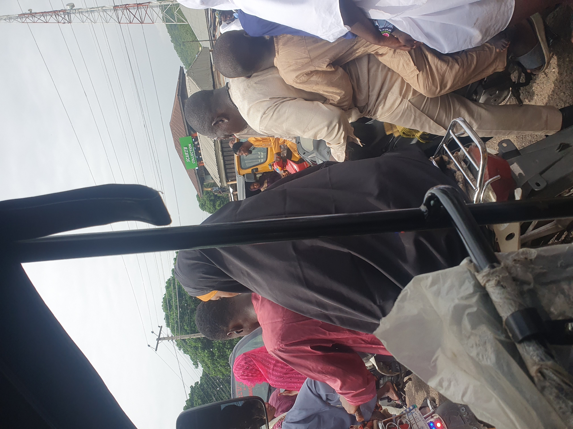 The image shows a crowded scene on what appears to be a motorcycle or tricycle in Lokoja, Nigeria. Several people are visible, including individuals wearing traditional attire such as a brown caftan and a black hijab. A yellow tractor is partially visible in the background, along with a building displaying a sign that says "SCAR2B COLLECTION". A red and white telecommunications tower stands in the distance. The view is taken from inside a vehicle, looking out.