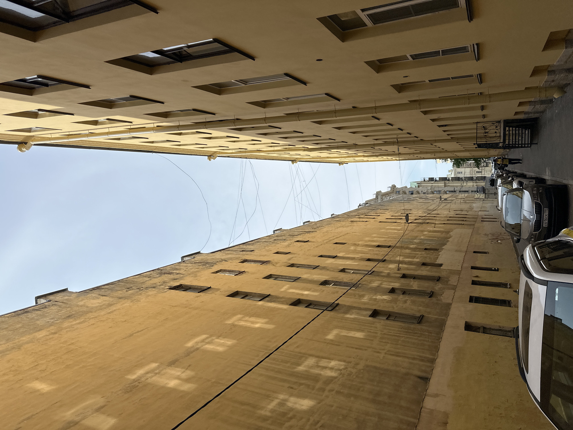 The image depicts a narrow courtyard or alleyway in Saint Petersburg, Russia, defined by two tall buildings with yellow facades. The buildings have numerous rectangular windows arranged in rows. Several cars are parked along the right side of the alley. A metal gate with decorative elements is also visible on the right side. Utility wires are strung between the buildings overhead. The sky is visible at the top of the frame.