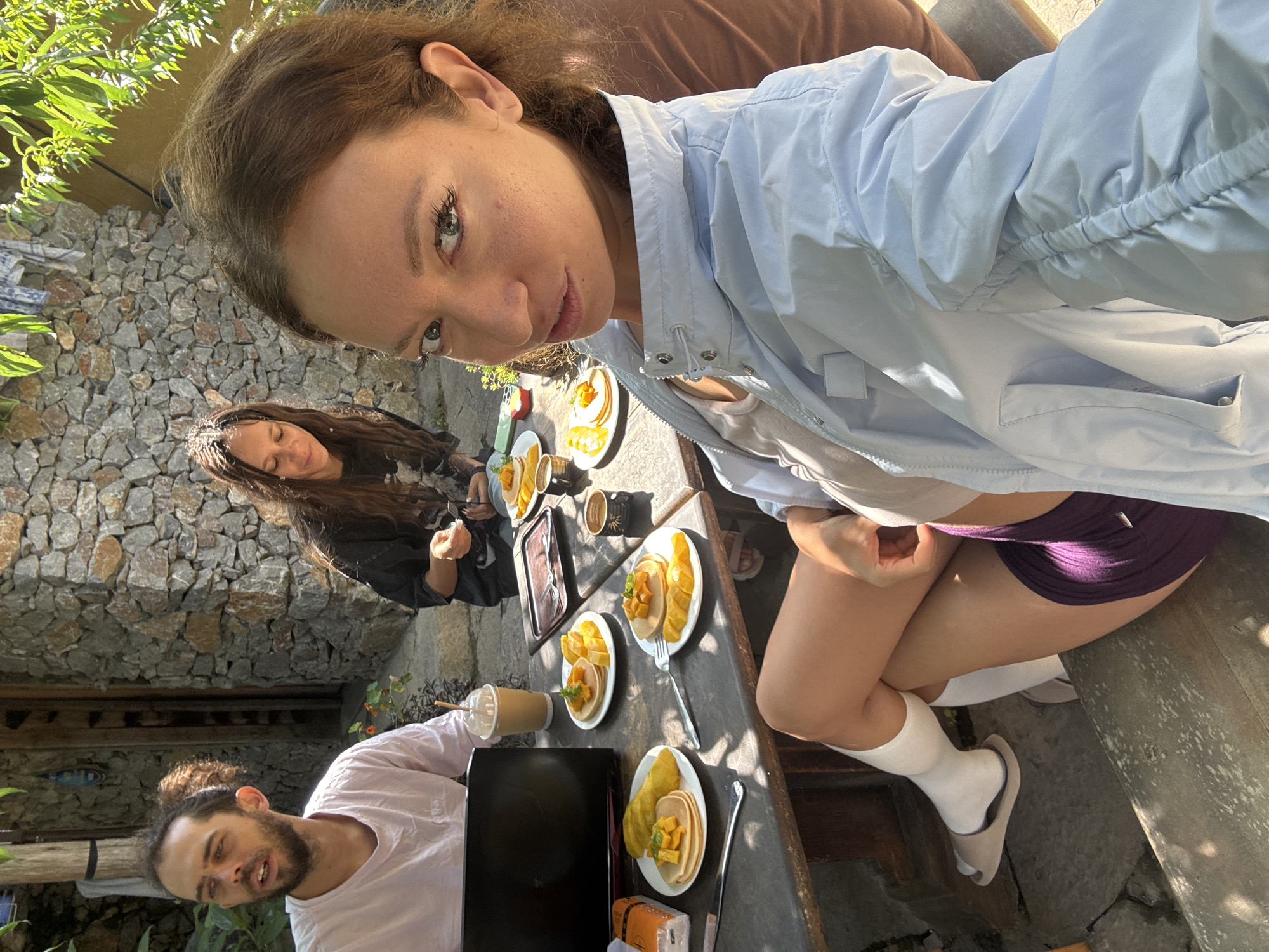 Three people are sitting at an outdoor table laden with plates of pancakes and fruit. A woman in a light blue jacket is taking a selfie. She is wearing a white top, purple shorts, white socks, and slides. Another woman is sitting behind the table, looking at her phone. A man is sitting at the other end of the table, using a laptop. The location appears to be an outdoor café or patio, possibly in Lũng Cú Commune, Vietnam, with a stone wall in the background.