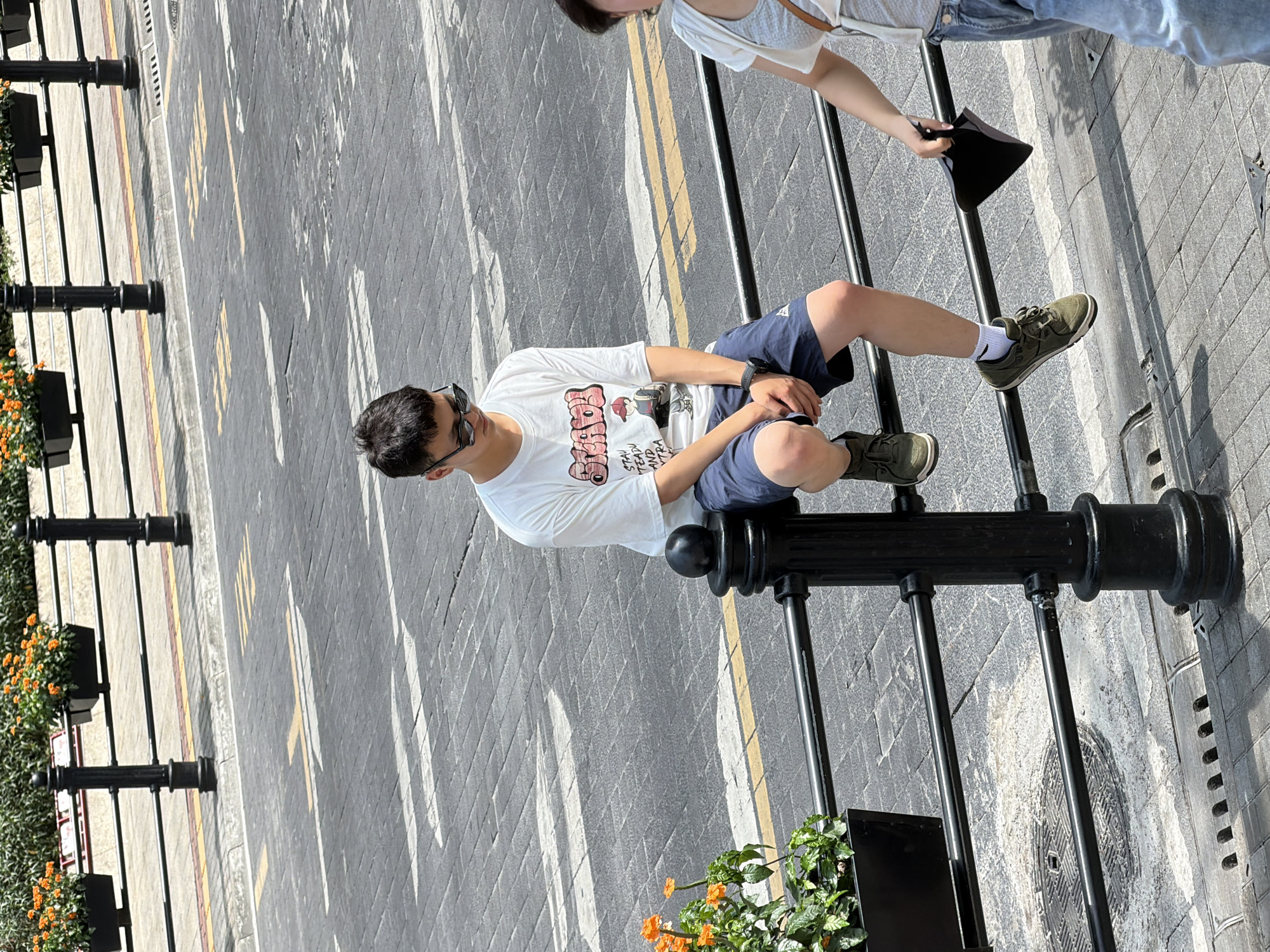 In Yuzhong District, China, a young man with short dark hair and sunglasses sits on a black metal railing. He is wearing a white t-shirt with a graphic print, dark blue shorts, and green sneakers with white socks. His left hand rests on his knee, and his right wrist has a watch. In the background, a person in a light-colored top and jeans walks away from the camera, holding a black object. The scene takes place on a street with a brick-patterned pavement and traffic markings. Orange flowers in black planters are visible along the railing.