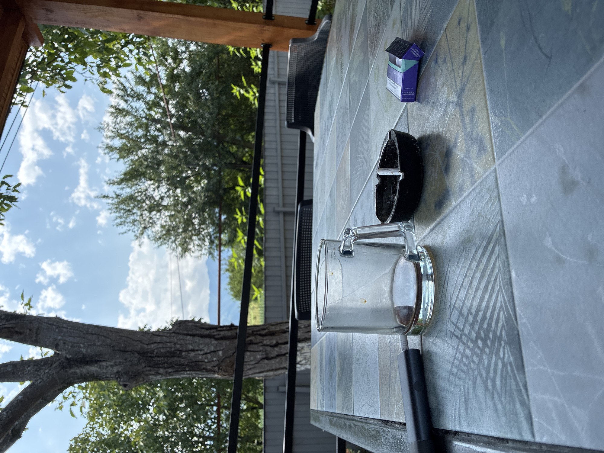 A glass mug with residual coffee sits on a tiled table outdoors, beside a black ashtray with a cigarette butt and a partially open pack of cigarettes. A black vape pen lies to the left of the mug. In the background, lush green trees and mountains are visible under a cloudy sky, suggesting a location in Surami, Georgia, possibly on a veranda or outdoor seating area.