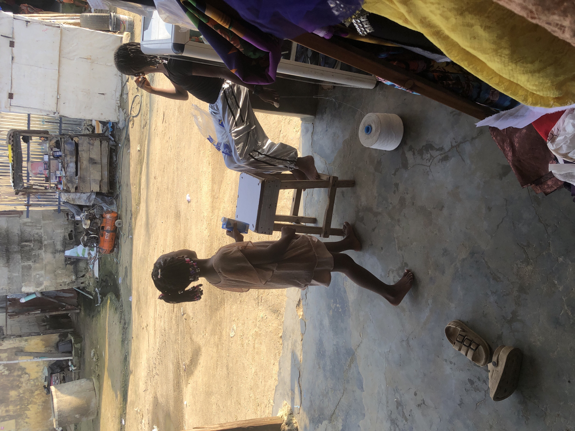 In Ado, Nigeria, a young girl in a light peach dress stands on a concrete floor, her back to the viewer. She holds a small blue object in her right hand and faces a small wooden stool with a white surface. To her right, a second person, wearing a black t-shirt and shiny silver pants, sits on the edge of a white plastic chair, facing away from the viewer and looking to the right. Their right hand is near their mouth. In the foreground, a pair of beige sandals lie on the concrete. Behind the seated person, a large spool of white thread rests on the ground. Further back, a wooden structure with shelves is visible, filled with various items. The background shows a dusty, unpaved area with several buildings constructed from various materials including concrete blocks and corrugated metal. An orange air compressor is parked to the left of a large concrete cylindrical structure. The overall scene depicts an outdoor, somewhat informal urban environment.