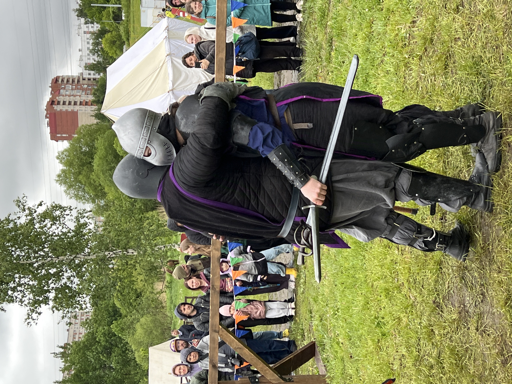 Two individuals in medieval armor are grappling in a grassy field. The individual on the left wears a grey helmet and dark quilted armor with purple trim. The individual on the right wears a darker helmet and full dark armor. The person on the left holds a sword, bracing it against the other's armor.
A crowd of people watches the event from behind a wooden barrier. Several children are among the spectators. Behind the crowd is a large white tent, a line of trees, and a multi-story apartment building.
The scene takes place in the city of Unknown, Unknown.