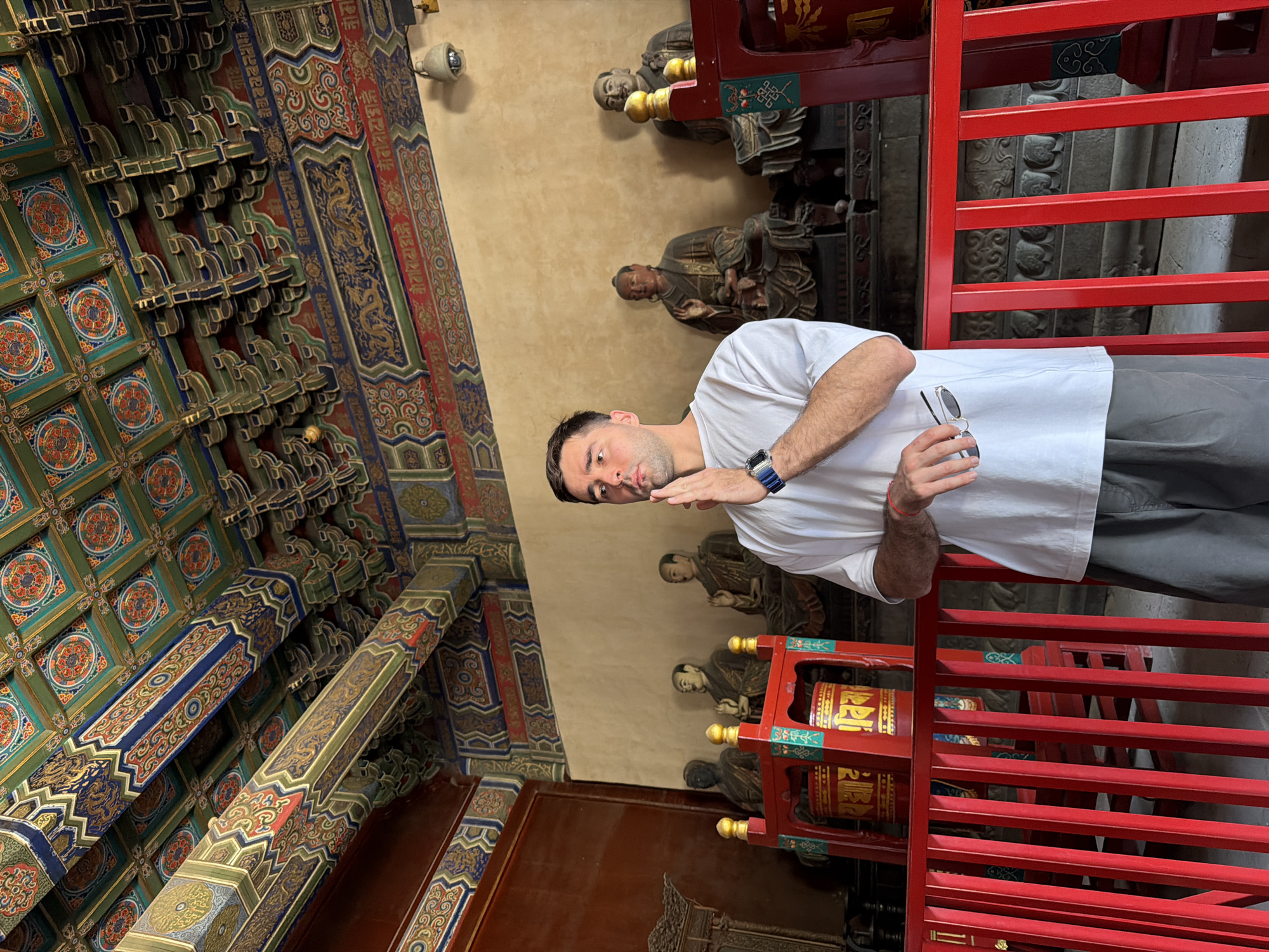 A man in a white t-shirt and grey pants stands inside what appears to be a temple or religious structure in the Chaoyang District of China, based on the architecture and artifacts. He is holding sunglasses in his hand, and his arm is raised as if he is gesturing or posing for a picture. The ceiling is elaborately decorated with green and blue patterns. There are wooden statues positioned along the wall behind him, as well as red structures that resemble prayer wheels or ceremonial objects.