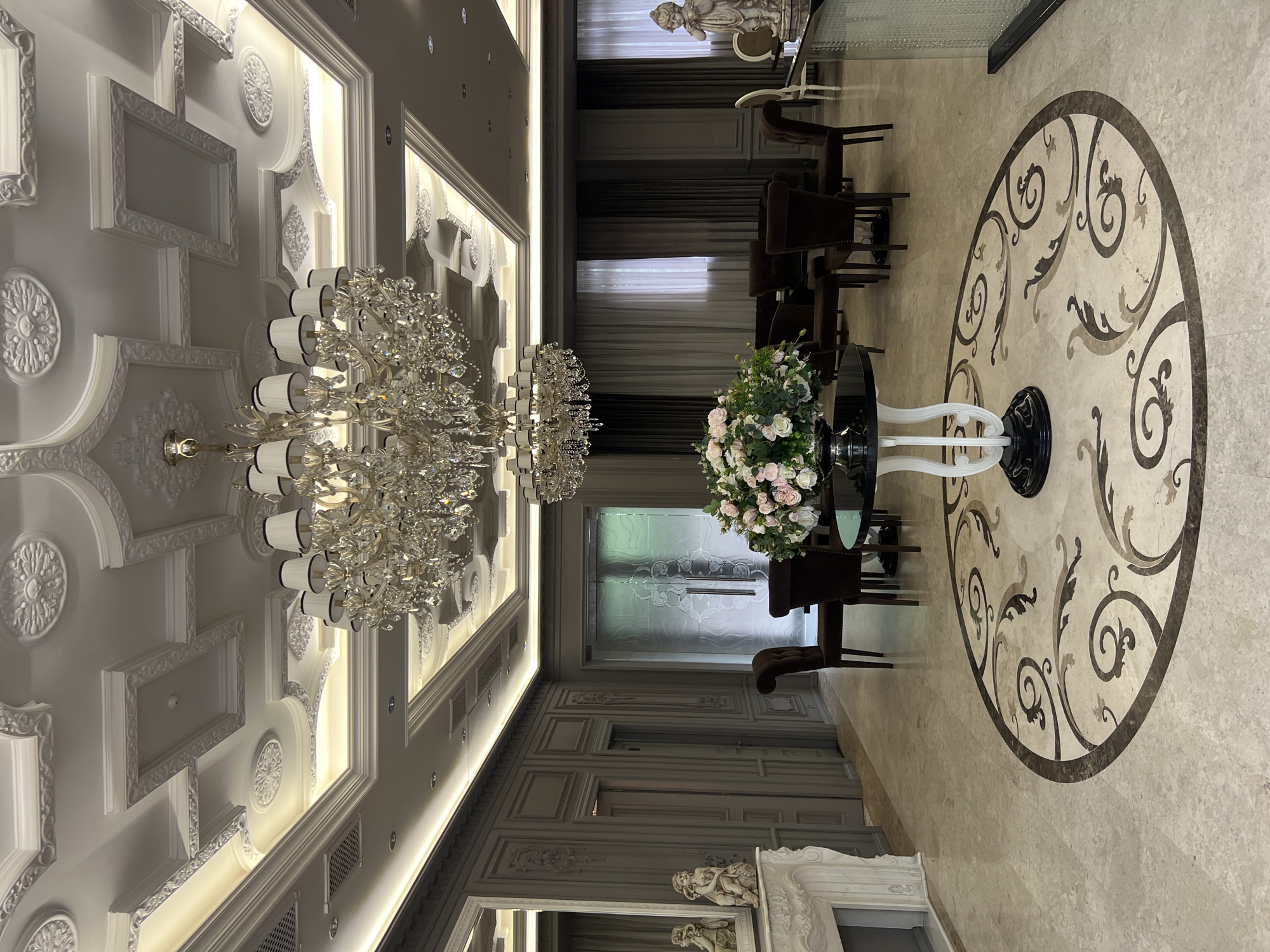 This image captures a luxurious and ornately decorated interior space, likely a grand hall or reception area, situated in Yerevan, Armenia. No people are visible.
The left side of the frame is dominated by a highly decorative ceiling featuring intricate white plaster moldings, including large circular medallions and rectangular frames. Warm LED strip lighting is recessed within coving along the ceiling's edges, complemented by scattered downlights. Two magnificent, multi-tiered crystal chandeliers, each adorned with countless sparkling prisms and elegant white cylindrical lampshades, hang from the ceiling, providing bright illumination.
The floor, visible on the right, is composed of polished light-toned marble or stone. A prominent circular mosaic inlay graces the center, showcasing an elaborate, swirling, baroque-inspired pattern in contrasting dark brown, lighter brown, and cream-colored marble.
Centrally positioned on the mosaic is a round black table with a distinctive white, sculptural pedestal base. It holds a large, lush floral arrangement overflowing with white and pink roses, peonies, and green foliage. Several dark brown, upholstered armchairs with wooden frames are arranged around this table, forming a seating area, with more chairs extending into the background.
The background of the room features dark, heavy curtains. Sections of a paneled wall with classical moldings are visible along the bottom of the image. A white, ornate structure, potentially a fireplace mantel or console table, adorned with classical carvings and topped by a small white statue, adds to the opulent decor. Another classical white statue rests on a dark surface in the upper background. A glass door with frosted, intricate patterns can be seen in the far distance.
The overall lighting is artificial and bright, creating a rich and sophisticated ambiance suitable for a high-end establishment. There is no visible text in the image.