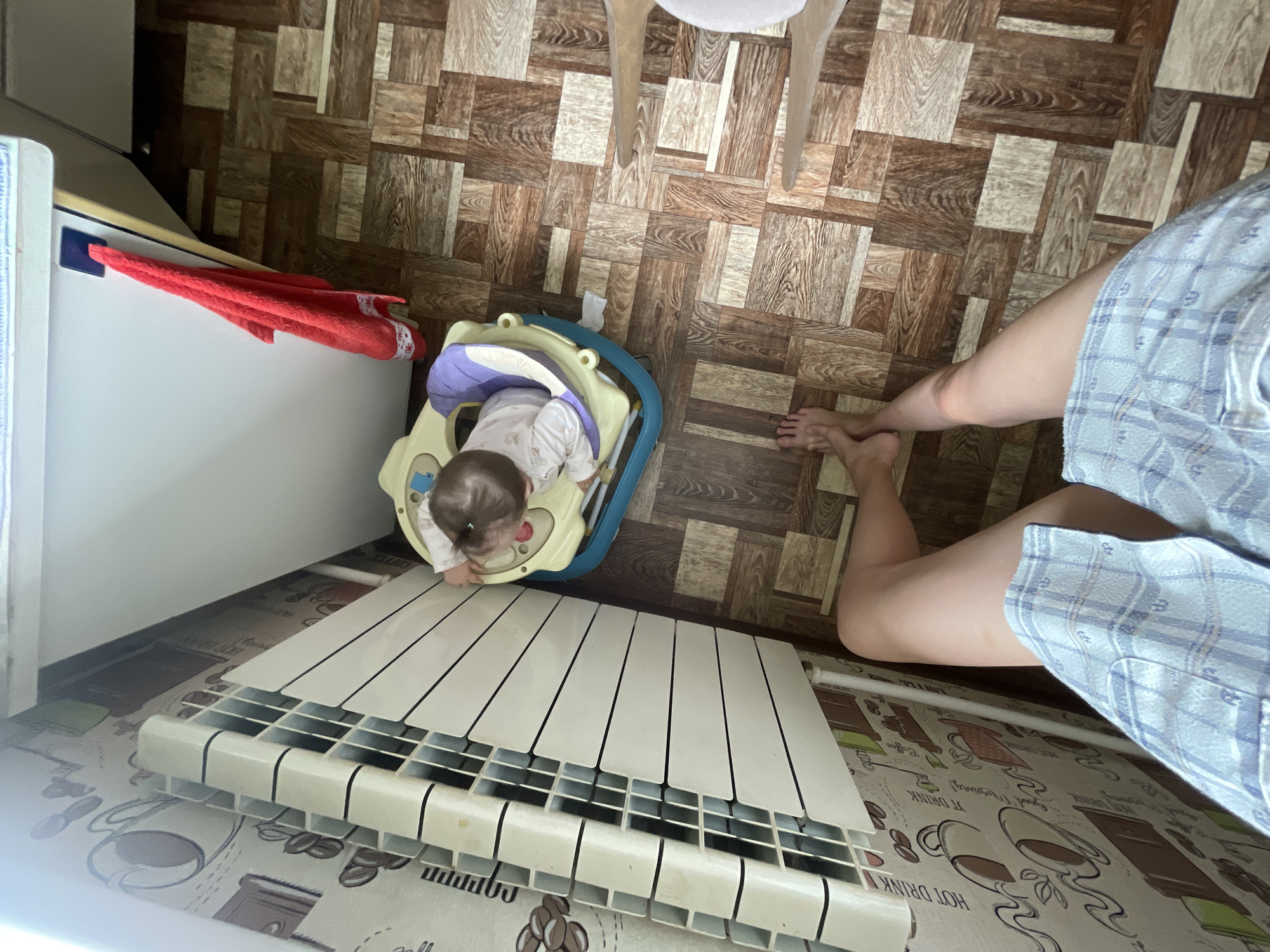 The image shows a top-down view of a domestic scene. A baby is in a walker, positioned near a radiator. A red towel is draped over a white appliance, possibly a refrigerator. The floor is tiled with a wood-grain pattern. The legs of an adult, wearing plaid shorts, are visible on the right side of the frame, suggesting the photographer is an adult. The scene appears to be indoors, likely in a home, given the presence of household items like the radiator, walker, and towel. There are no strong indicators of time of day or weather conditions. The overall mood is casual and domestic. Visible text on a rug beneath the radiator includes "HOT DRINK" and other decorative elements like coffee cups and beans. The location context is Sharkan, Russia.
