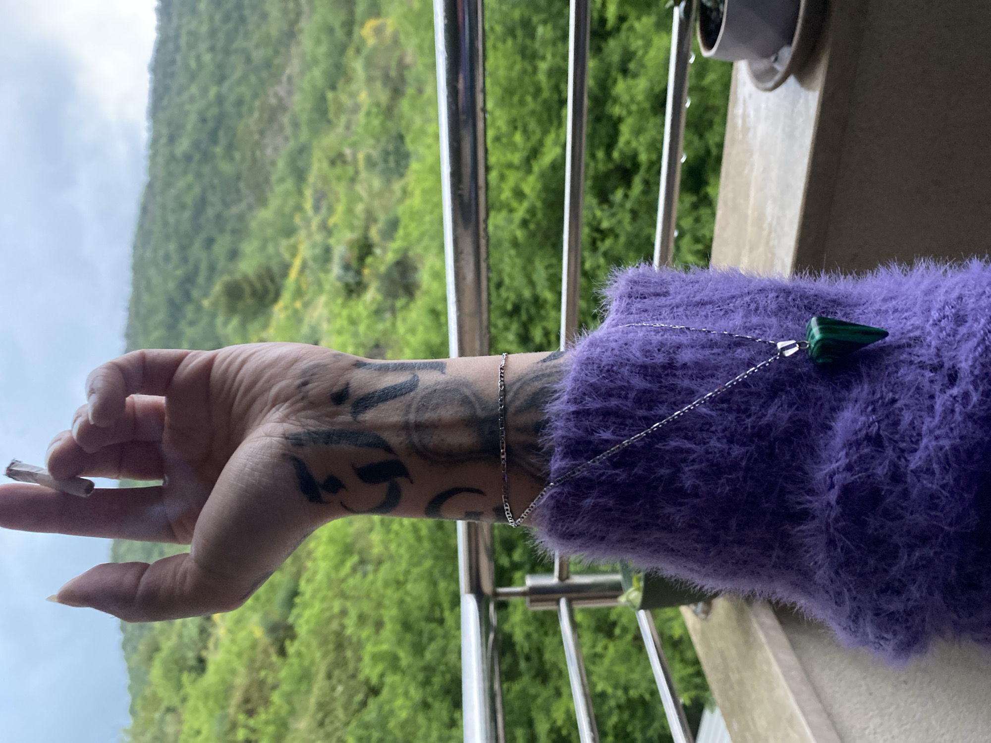 A person with tattoos on their arm is holding a lit cigarette, and wearing a purple sweater with a green pendant necklace. They are standing on a balcony overlooking a mountain range in Boreti, Montenegro. The sky is overcast.