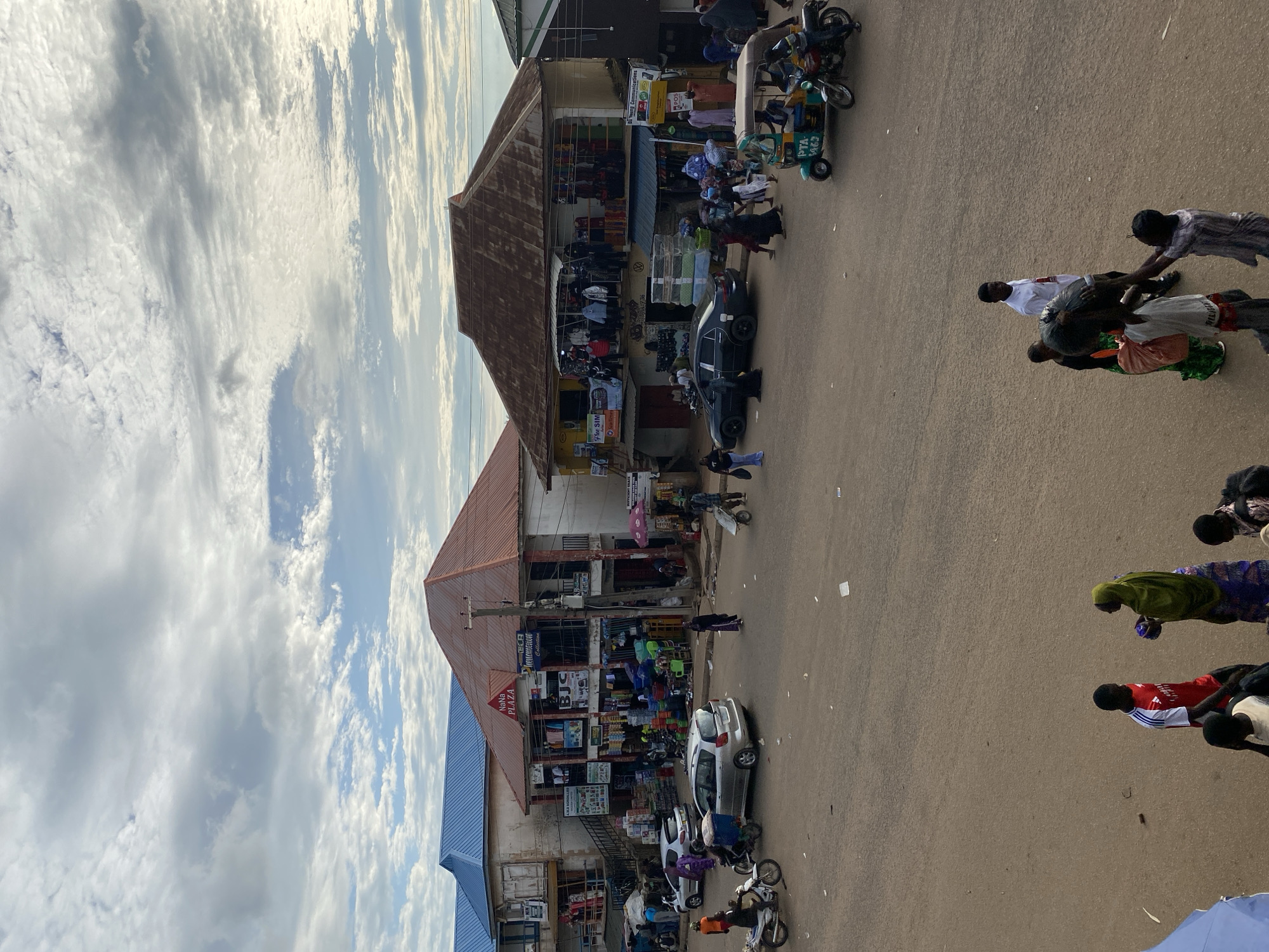 This image depicts a bustling street scene in Bukuru, Nigeria, under a predominantly cloudy sky with patches of blue. The time appears to be daytime, with soft, diffused natural light.
The scene is dominated by a wide, unpaved or dirt-covered road that curves slightly through the center, with multiple two-story commercial buildings lining both sides. The architecture of these buildings varies, but most feature corrugated metal roofs in shades of blue, rusty red, and dark brown. Many buildings house shops and businesses, indicated by goods displayed outside and numerous signs. Visible signs include "NANA PLAZA," "BJC," "A.M.D Agricultural," "FREE SIM," "StarTimes," "Young Communications," and "POS." Utility poles laden with power lines are prominent along the street, particularly in the middle ground.
Numerous people are present, engaging in various activities typical of a market or commercial area. Individuals are seen walking across and along the street, some carrying goods on their heads or in bags. One person pushes a two-wheeled handcart. Several vehicles are on the road: a silver hatchback (possibly a Toyota Yaris), a white hatchback, and a black sedan (possibly a Mercedes-Benz C-Class). Multiple motorcycles are also present, one with a rider and a large black bag, and another with a passenger. A light blue and green motorized tricycle (Keke NAPEP) labeled "PTA M63" is seen with passengers.
In the foreground, a group of people stands on the dirt road. One man wears a red and white striped shirt (resembling an Arsenal football club jersey). Another woman, dressed in a green headscarf and a patterned skirt, holds a blue object. Further to the right in the foreground, a man in a white shirt is visible, along with others carrying bags. The overall impression is one of active commerce and daily life in an urban Nigerian environment.