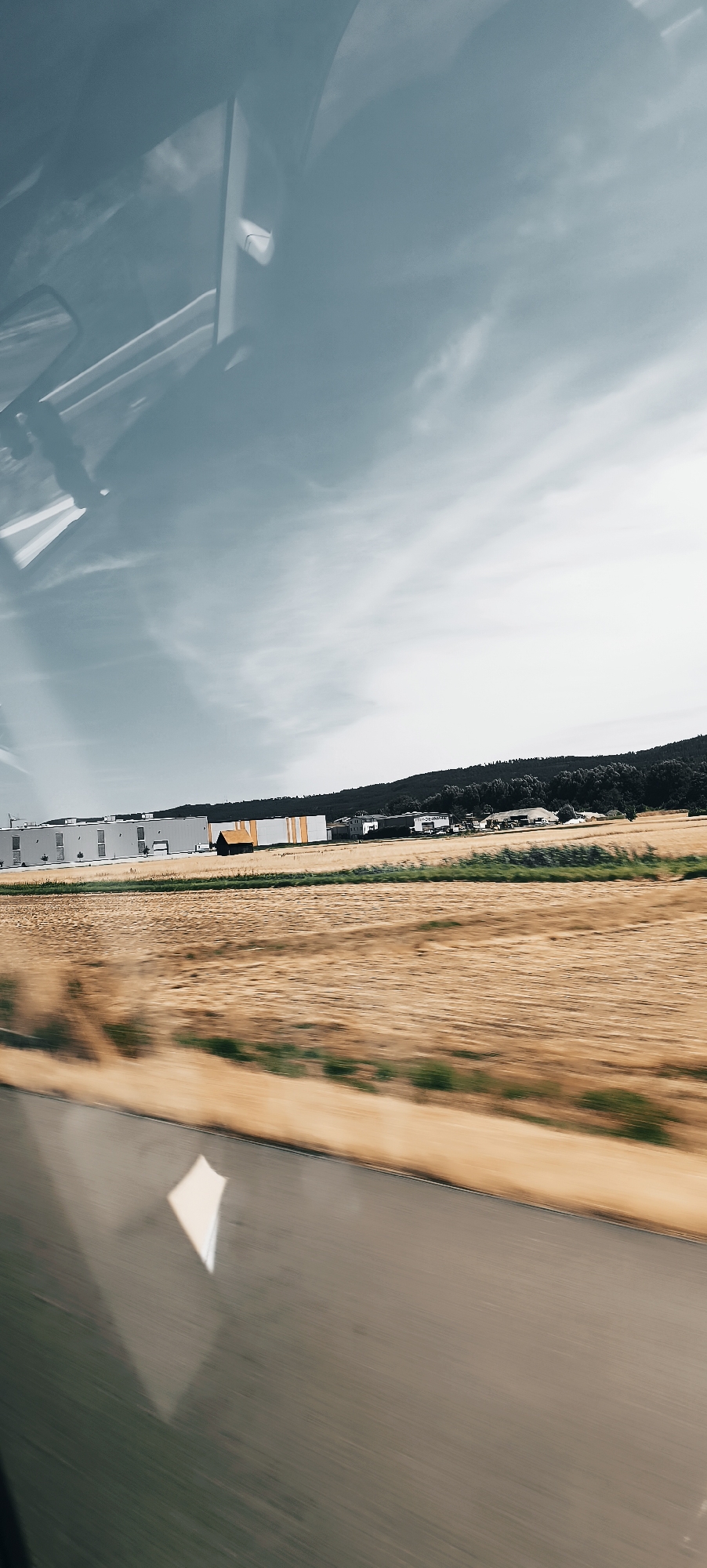 The image captures a blurred outdoor scene, likely taken from a moving vehicle. In the foreground, a grey, blurred surface resembling a road is visible. Above the road, a field of dry, golden grass is out of focus. Further in the background, a long, low-slung building complex with a white facade and orange accents is situated. To the right of the complex, a small, dark, gable-roofed structure stands in a field. Behind the buildings, a dark line of trees forms a hill. The sky above is a pale blue with wispy clouds. Reflections of the vehicle's interior, including a white structure and parts of the windshield, are visible in the upper portion of the image. The scene is likely in the vicinity of Winterbach, Germany, given the industrial architecture and rural landscape.