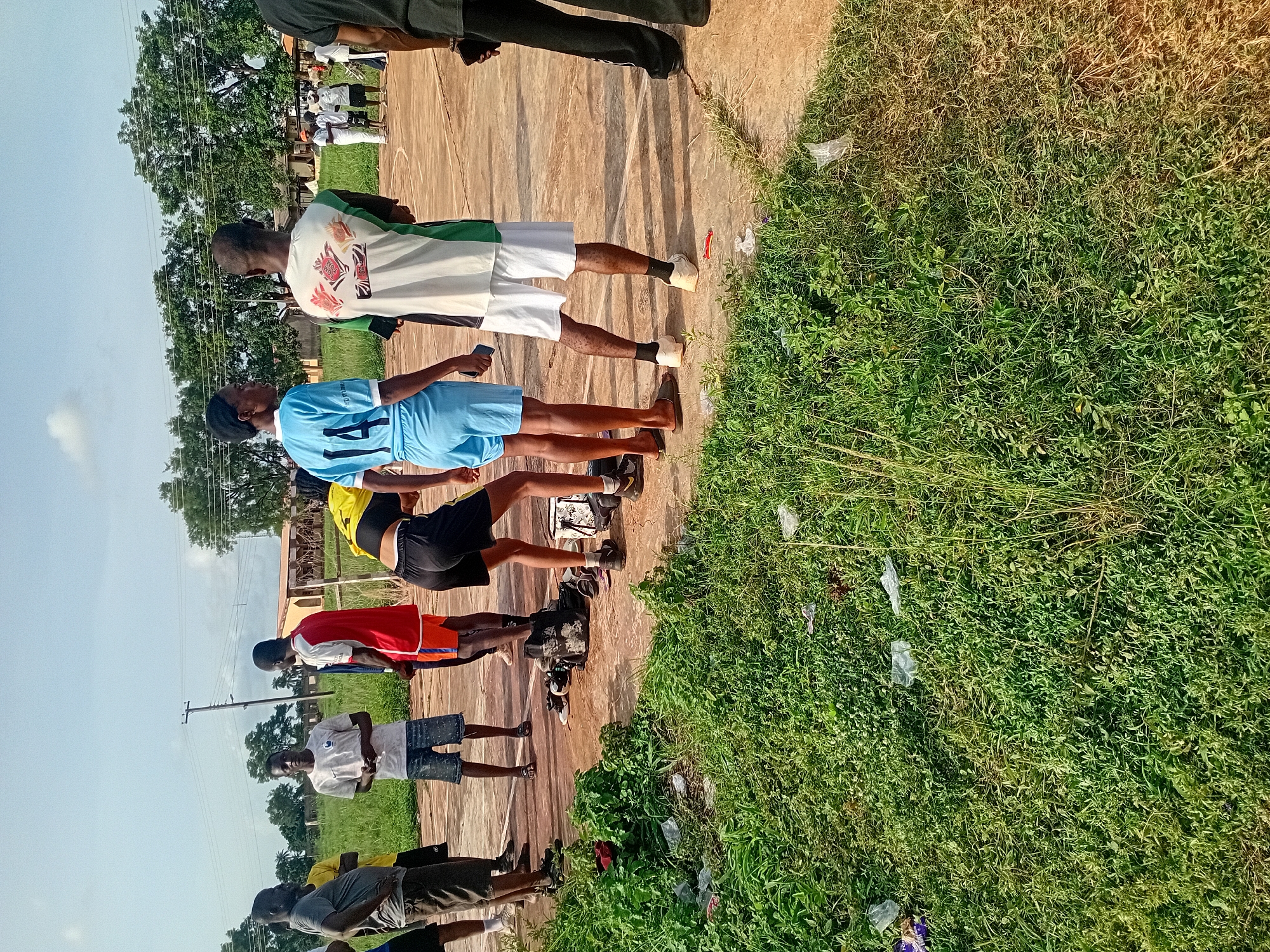A group of young men are standing on a dirt patch in Ogbogbo, Nigeria, some wearing jerseys, with a grassy area beside them, under a clear sky. Some garbage is scattered in the grass.
