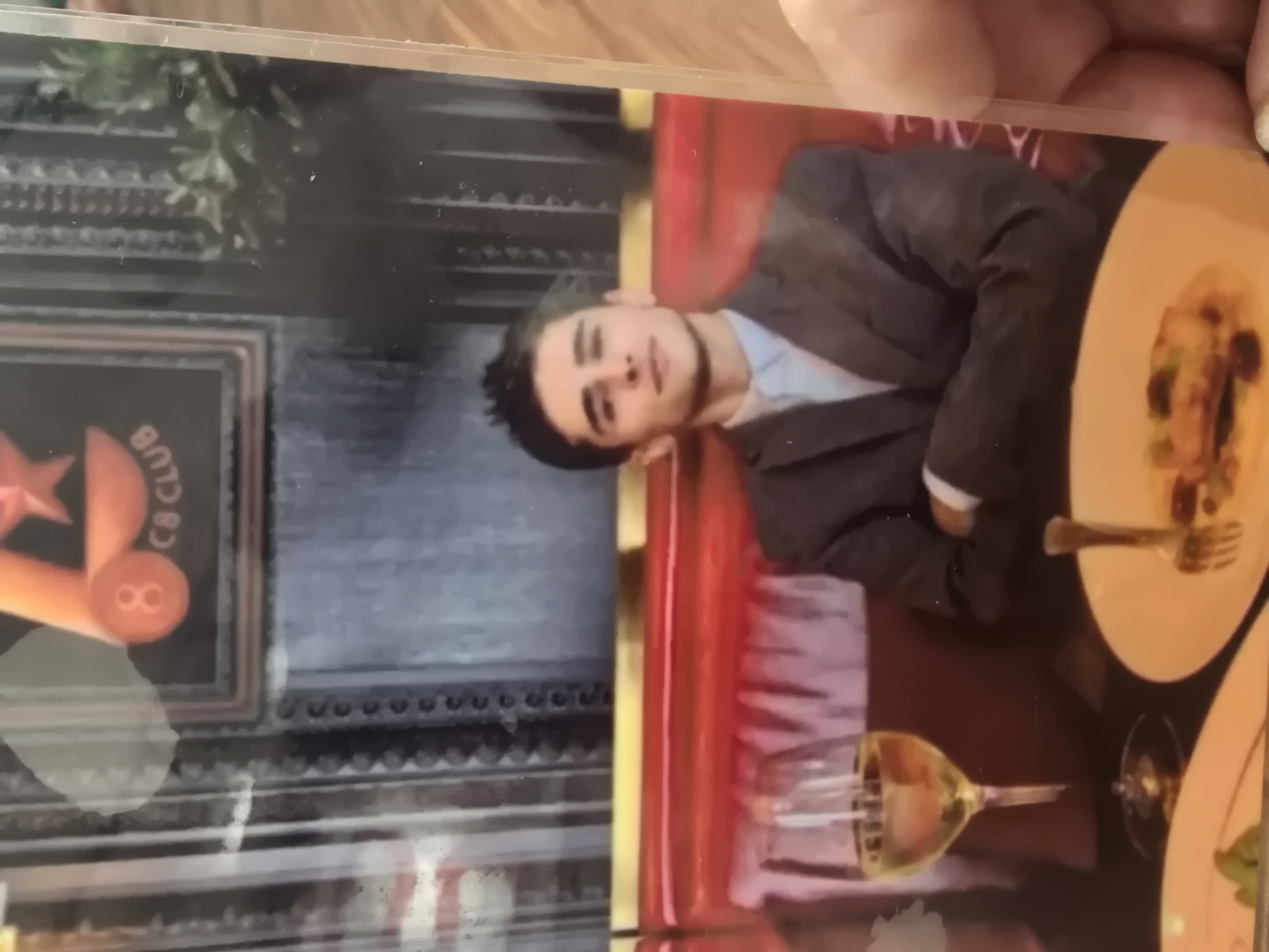 The image depicts a young man dressed in a suit and light blue shirt, seated at a table in what appears to be a restaurant or club. He is looking towards the camera with a neutral expression. In front of him is a glass of white wine and a plate with food and a fork. The background shows red upholstered seating and a framed artwork on the wall with the text "8 C8 CLUB". The scene is set indoors, with diffused lighting. There is no visible indication that this location is in Ulan-Ude, Russia.