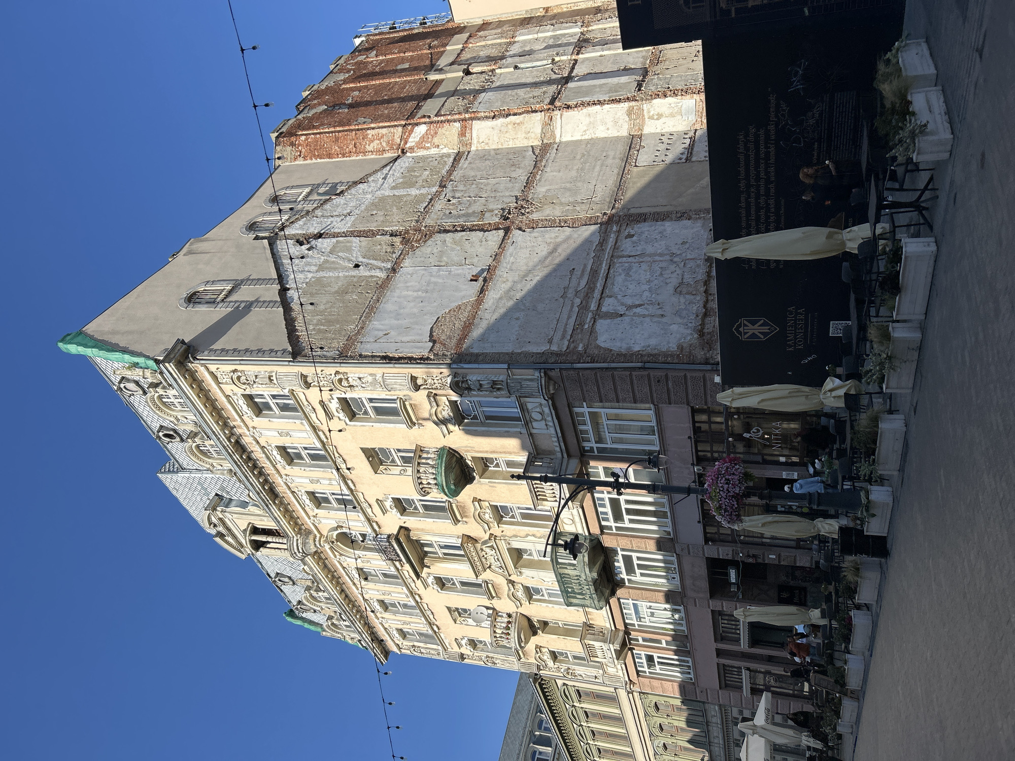 The image shows a multi-story building on a city street. The building is partially under renovation, with one side of the facade removed, exposing brickwork and concrete. The remaining part of the building has a classical architectural style with decorative elements, balconies, and mansard roofs. In front of the building, there is a cafe or restaurant area with tables, chairs, and umbrellas set up on a paved walkway. Several people are visible, some seated at tables and others walking by. String lights are suspended across the street. The sky is clear and blue. The building is located at Piotrkowska 69 in Łódź, Poland, as indicated by signage.