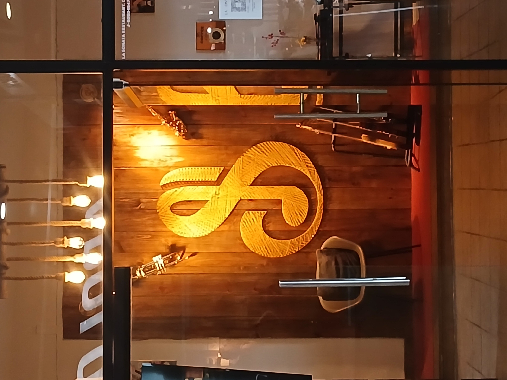 The image shows a window display of "La Sonata Restaurant" in Guayana City, Venezuela. The interior is decorated with wooden planks on the wall, featuring a large, intertwined musical note symbol made of light-colored material. There are musical instruments displayed, including what appears to be a saxophone and other brass instruments. A chair is visible within the display. A string of decorative lights hangs to the left of the display. The restaurant's name and possibly a phone number are visible in the top section of the window.