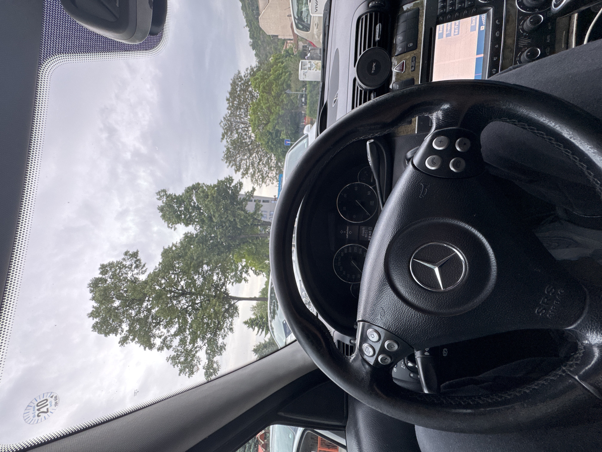 The image appears to be taken from inside a Mercedes-Benz vehicle, looking through the windshield. The car's dashboard and steering wheel, featuring the Mercedes-Benz logo, are prominent in the foreground. Outside the car, trees and buildings can be seen. The image also shows a "TÜV SÜD" sticker on the windshield, indicating a vehicle inspection. Based on the presence of a "VIVA" branded vehicle, the location is possibly Plovdiv, Bulgaria. The image captures a view from inside the car towards a street with other vehicles and a cloudy sky.