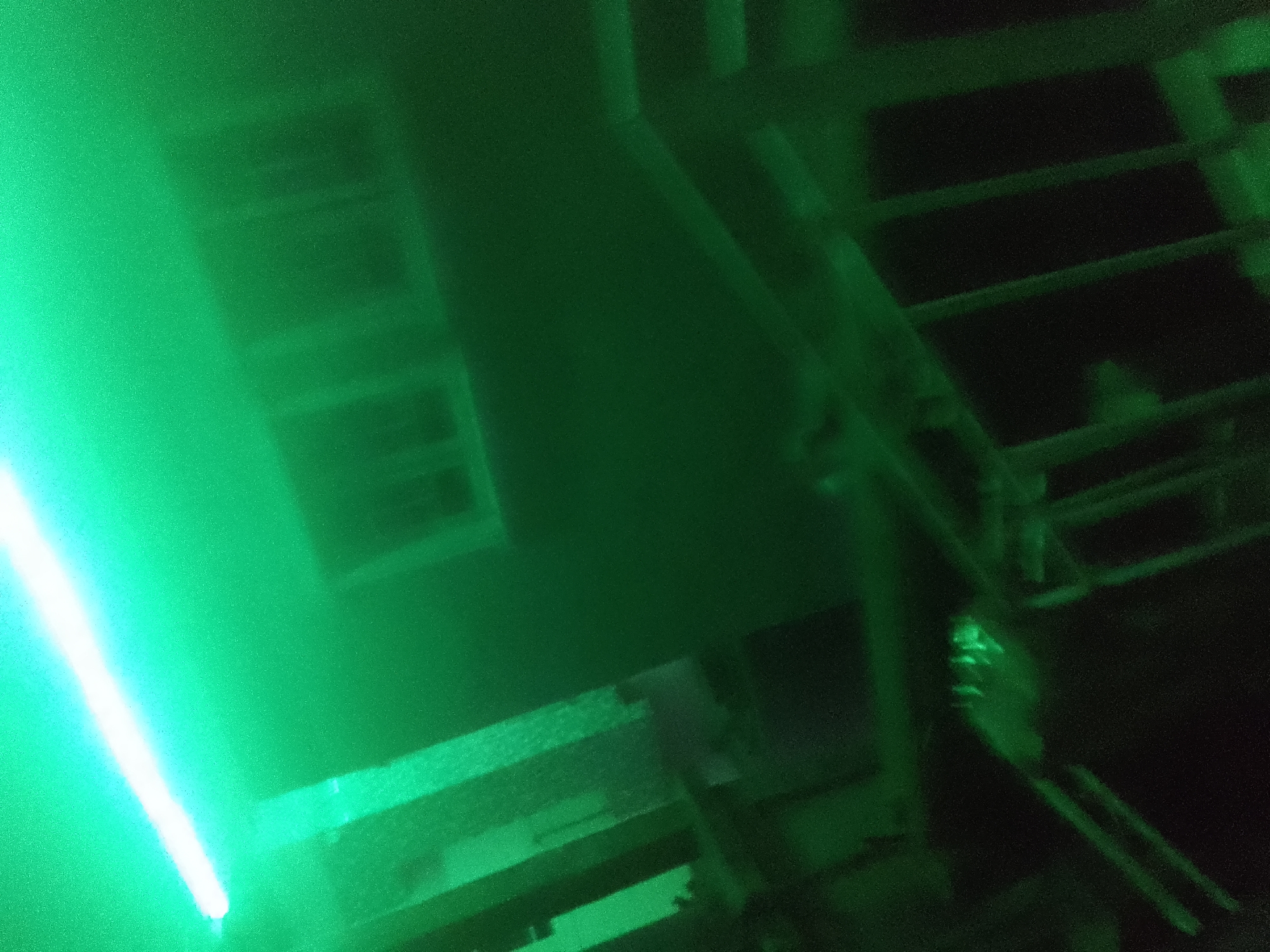 The image, taken in Jalingo, Nigeria, appears to be an interior shot illuminated by a bright green light source, possibly a laser or LED light, casting a strong green hue across the scene. In the foreground, a bright, vertical beam of green light is visible on the left side of the frame. The background shows indistinct shapes of what could be windows or openings in a wall, with dark, horizontal structures that suggest shelving or a tiered arrangement of objects. The overall impression is one of a dimly lit interior space, possibly a workshop, storage area, or an industrial setting, heavily influenced by the intense green lighting. The image is grainy and out of focus, making precise identification of objects challenging.