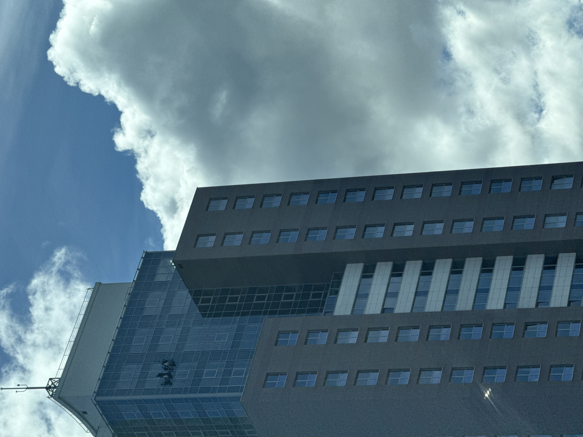 The image shows a close-up, low-angle shot of a tall, modern skyscraper with a glass facade on its upper section and a darker, more solid facade on its lower section. Several window cleaners are suspended by ropes on the glass section of the building, cleaning the windows. The building is silhouetted against a bright blue sky filled with dramatic, white clouds. Part of the building appears to be under construction or undergoing renovation, as indicated by the presence of workers on the facade. The architecture suggests a contemporary urban environment. This skyscraper is likely located in Riga, Latvia, given the style of architecture and the presence of high-rise buildings in the city.