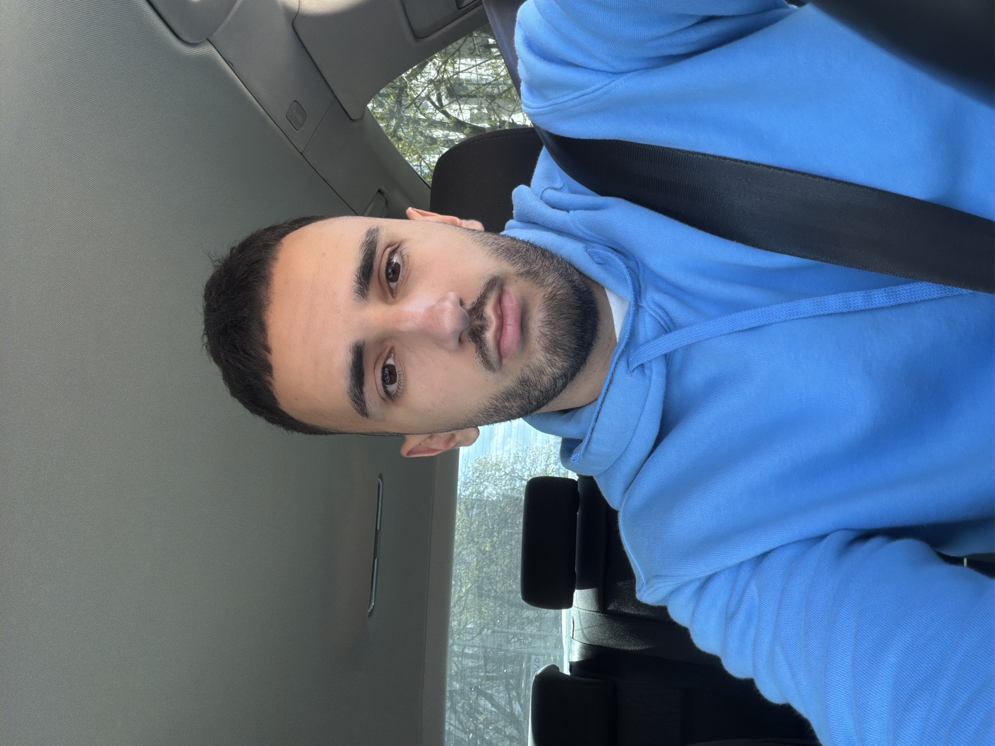 A man with dark hair and a beard is sitting in a car. He is wearing a light blue hooded sweatshirt and a black seatbelt. The interior of the car is visible, including the headrest of the seat behind him. The background outside the car window is blurry, but appears to show trees. There is no verifiable evidence within the image to place the location in the city of Plovdiv, Bulgaria.