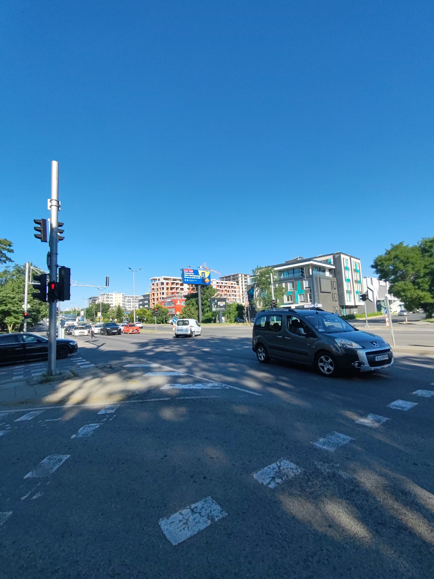 The image depicts a street intersection in Burgas, Bulgaria, under a clear blue sky. A traffic light with a red signal for vehicles is prominently visible on the left, illuminating a pedestrian crossing. Several cars are stopped at the intersection, including a black sedan on the left and a gray minivan driving through the intersection from the right. In the background, modern residential buildings with distinctive green accents are under construction, along with other completed buildings. A large billboard advertisement is also visible. The street is paved with asphalt, and traffic markings, including pedestrian crossings and lane dividers, are clearly visible. Trees line the sides of the street, and the overall scene suggests a typical urban environment on a sunny day.