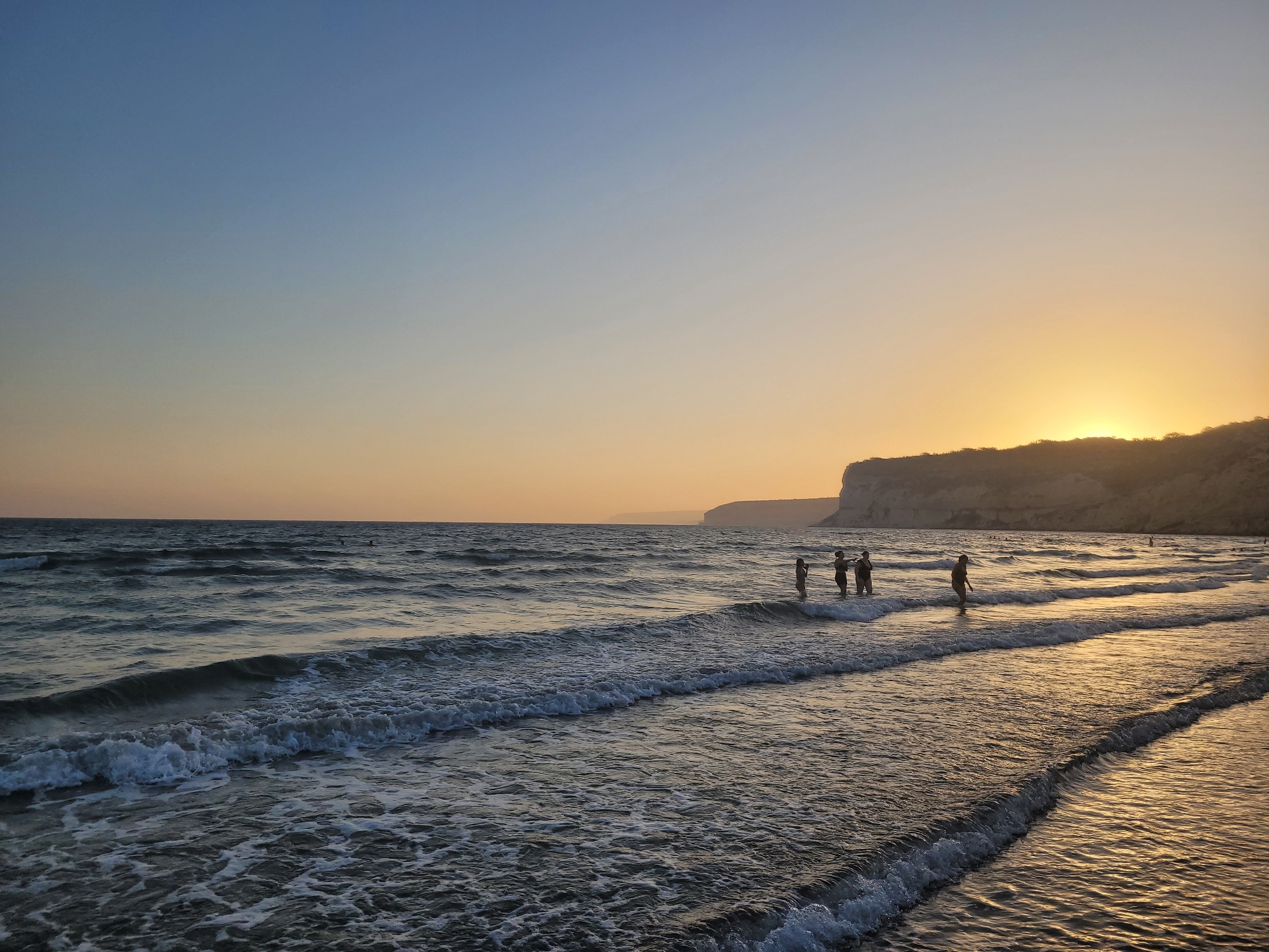 The image depicts a serene beach scene at dusk. The sky transitions from a clear blue at the top to a warm orange hue near the horizon, where the sun is setting behind a distant cliff. Gentle waves roll onto the sandy shore, reflecting the golden light of the sunset. Several people are visible in the shallow water, some standing and others wading, appearing to enjoy the calm evening. The environment suggests a peaceful, natural setting, likely a coastal area. The reference to Episkopi Lemesou, United Kingdom, appears to be an error, as Episkopi is a village in Cyprus.
