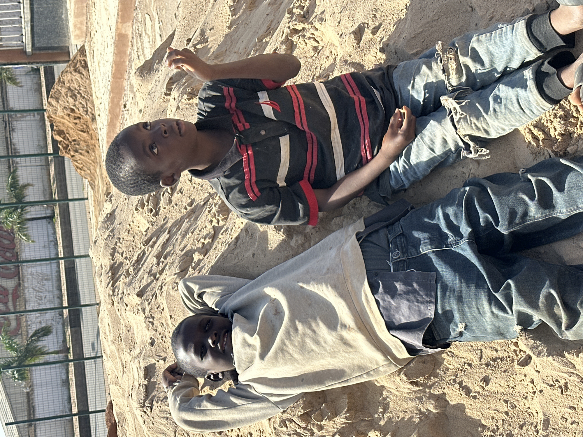 Two young boys are sitting on a pile of sand. The boy on the left is lying down with his hands behind his head. He is wearing a light-colored long-sleeved shirt and blue jeans. The boy on the right is sitting upright and looking upwards, holding a small amount of sand in his right hand. He is wearing a striped polo shirt and distressed blue jeans. In the background, there is a fence, some palm trees, and a building with a Coca-Cola logo. The scene appears to be an outdoor construction or play area in Jos, Nigeria.