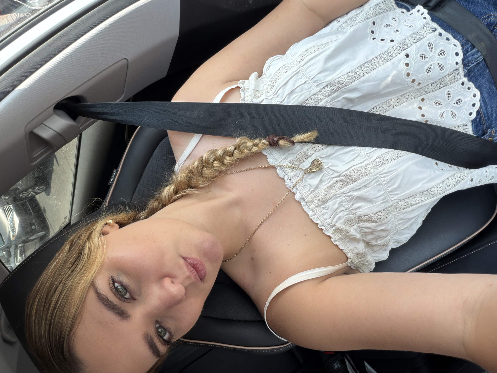 The image is a high-angle, close-up selfie of a young woman seated inside a car, likely in Puebla, Mexico, during the daytime. She has fair skin, and her light blonde hair is styled in a single braid extending over her left shoulder, secured with a brown elastic band. Her striking green eyes look directly at the viewer, framed by defined eyebrows and long eyelashes. She wears a white top featuring intricate eyelet embroidery and scalloped edges, with thin shoulder straps. A delicate gold chain necklace with a small circular pendant adorns her neck. A black seatbelt is visible across her chest and lap, indicating she is buckled in. Parts of her blue denim jeans are visible on her right side. The car's interior includes a dark, perforated leather seat with the word "AIRBAG" clearly visible on its side bolster, and a grey plastic trim piece by the window. Bright natural light illuminates the scene, suggesting a sunny day. The woman maintains a serene and direct gaze, conveying a sense of calm.