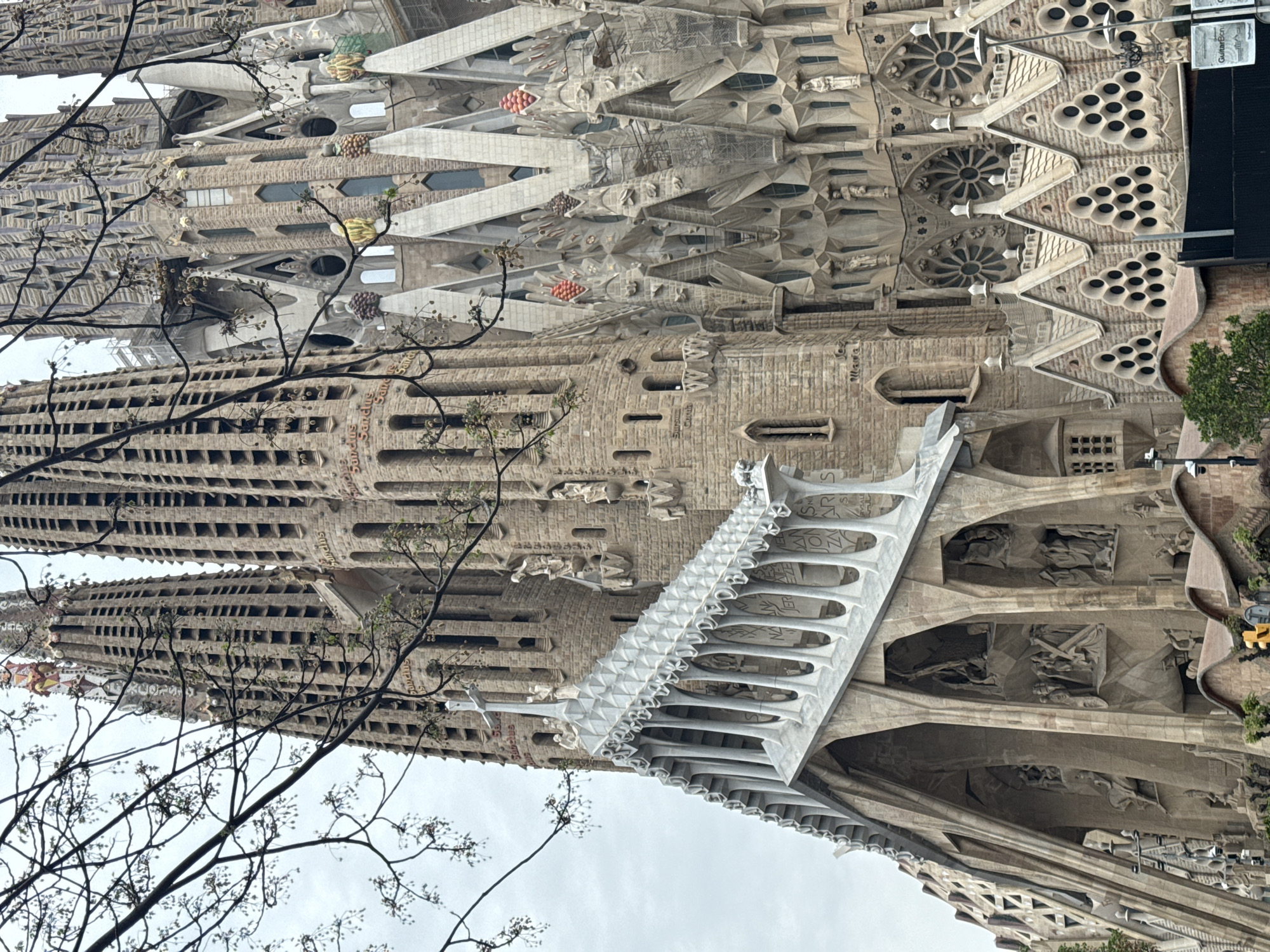 The image shows a partial view of the Sagrada Familia, a large unfinished Roman Catholic minor basilica in Barcelona, Spain. The basilica's facade is characterized by its intricate, highly ornamented design, including spires, windows, and sculpted details. A white structure, presumably a bridge or walkway, juts out from the building. The sky is overcast. A bare tree with sparse foliage is partially visible in the foreground.