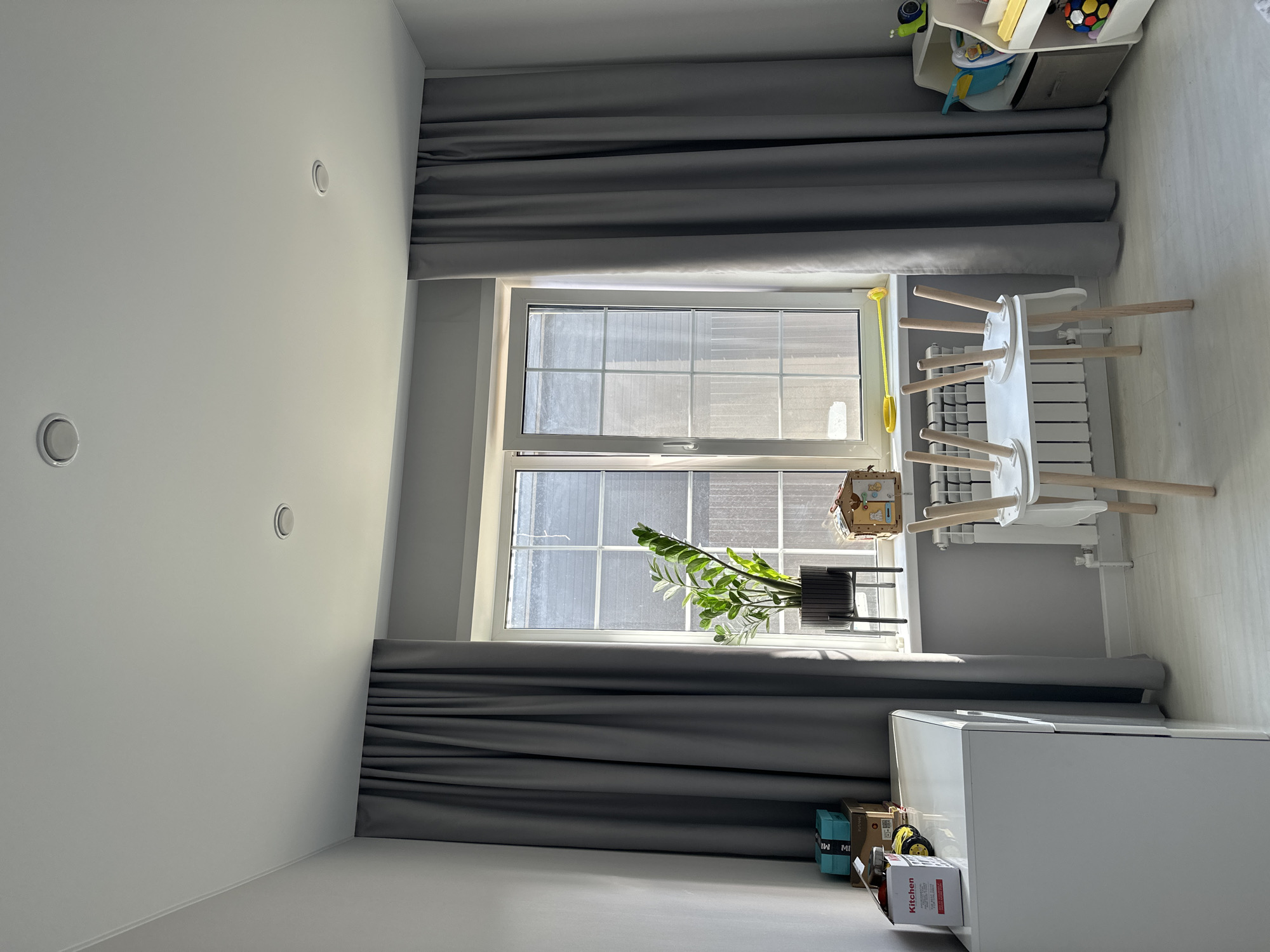 In a room with light gray walls and ceiling, a large white dresser is on the left. On top of the dresser are several boxes, including one labeled "Kitchen". To the right of the dresser, gray curtains frame a window. A potted plant with large green leaves sits on the windowsill. A small wooden house-shaped object is also on the windowsill, next to the plant. In front of the window is a white table with wooden legs, currently upside down. To the right of the table is a white toy organizer with various toys, including a ball and a toy snail. A white radiator is visible beneath the window. The floor is light-colored wood. The room appears to be a child's bedroom or play area. The image was taken in the city of Estachi, Russia.