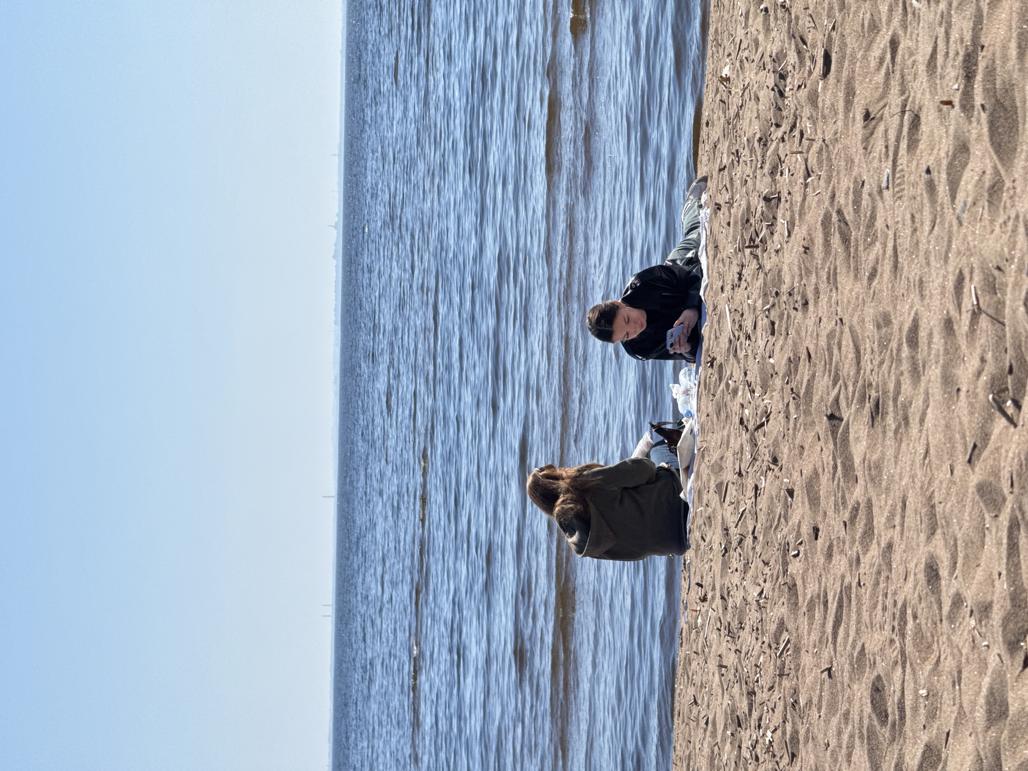 Two people are sitting on a sandy beach. One person is wearing a black jacket and looking at their phone. The other person is wearing a green jacket with a fur hood and looking towards the water. The location is Komarovo, Russia, judging from the landscape, with a body of water and a distant cityscape visible in the background. There is a bottle of water and a plastic bag near the person with the black jacket. The beach sand is textured and contains small debris like twigs.