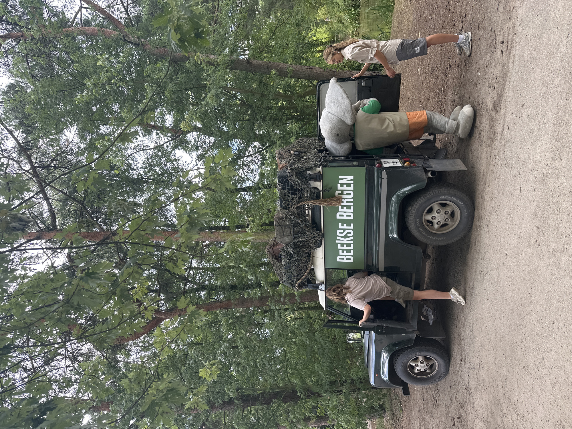 A dark green, open-door Land Rover Defender is parked on a dirt path. The vehicle has a camouflage net covering the roof rack. On the side of the Defender, the text "BEEKSE BERGEN" is visible in white capital letters. A person with blonde hair, wearing a light brown t-shirt and khaki shorts, is climbing into the driver's side of the vehicle. Facing the vehicle from the passenger side is another blonde person wearing a light brown collared shirt and dark grey shorts, holding the open rear door. Standing beside the rear of the vehicle is a person in a grey elephant costume with a green vest and orange shorts. The background is a dense forest with tall trees. In the far right background, a body of water can be seen. The overall scene appears to be at Beekse Bergen Safari Park in Hilvarenbeek, Netherlands, given the text on the vehicle.