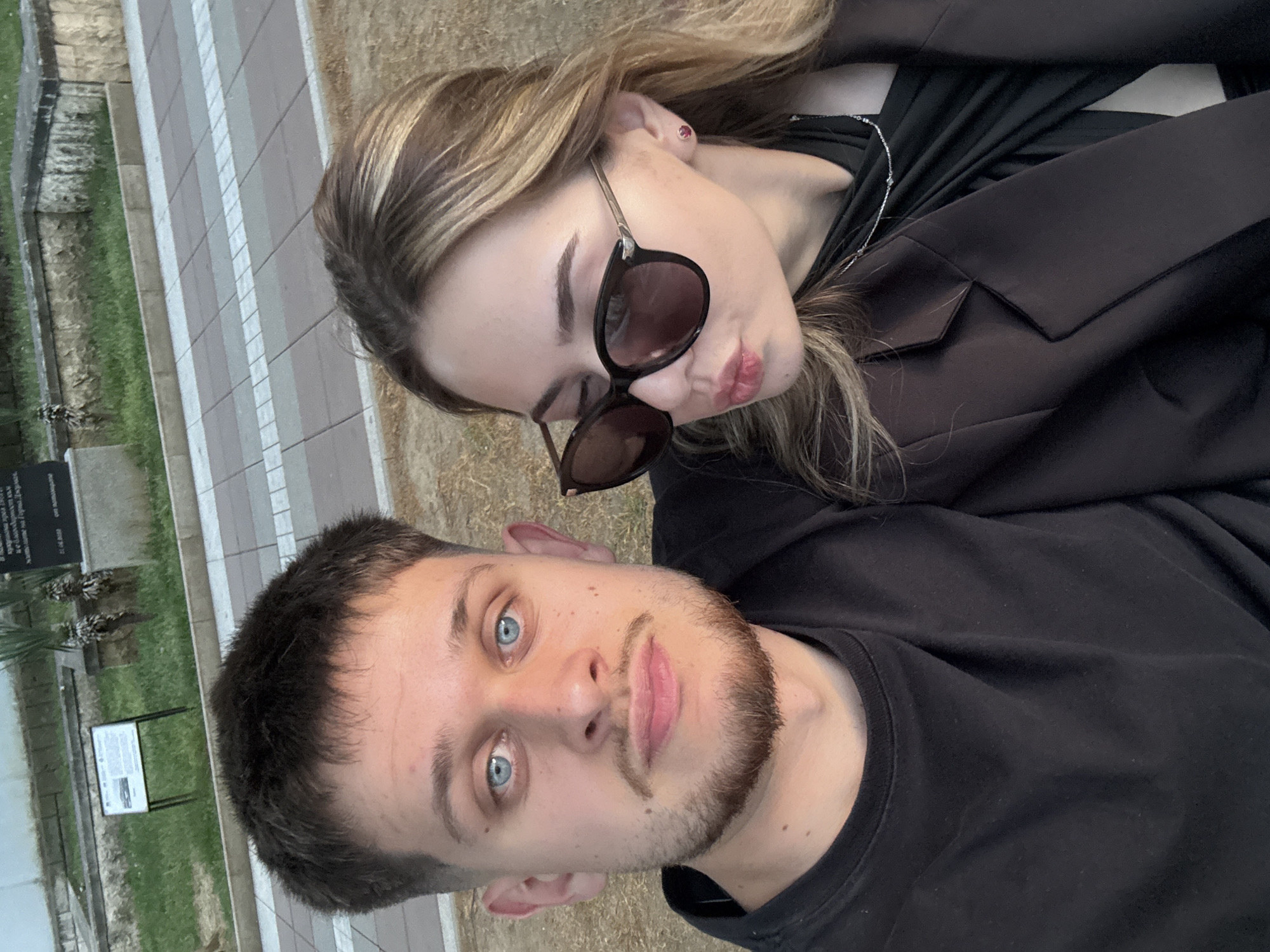 A close-up selfie shows a man and woman posing, with the man in the foreground and the woman slightly behind him. The man has fair skin, blue eyes, and short dark hair with a trimmed beard. He wears a black t-shirt. The woman, positioned above and behind him, has light brown hair with blonde highlights. She wears sunglasses and has on a dark jacket over a dark top with a necklace.
The background includes a paved area with a distinctive pattern of white and gray rectangular tiles, and a patch of grass with a dark memorial plaque and informational sign. Based on the visual cues of the surroundings, the picture appears to be taken in the city of Blagoevgrad, Bulgaria, near a historical or commemorative site, where the paving and informational signage are common features. The angle suggests the picture was taken with the camera positioned above the subjects, looking down.