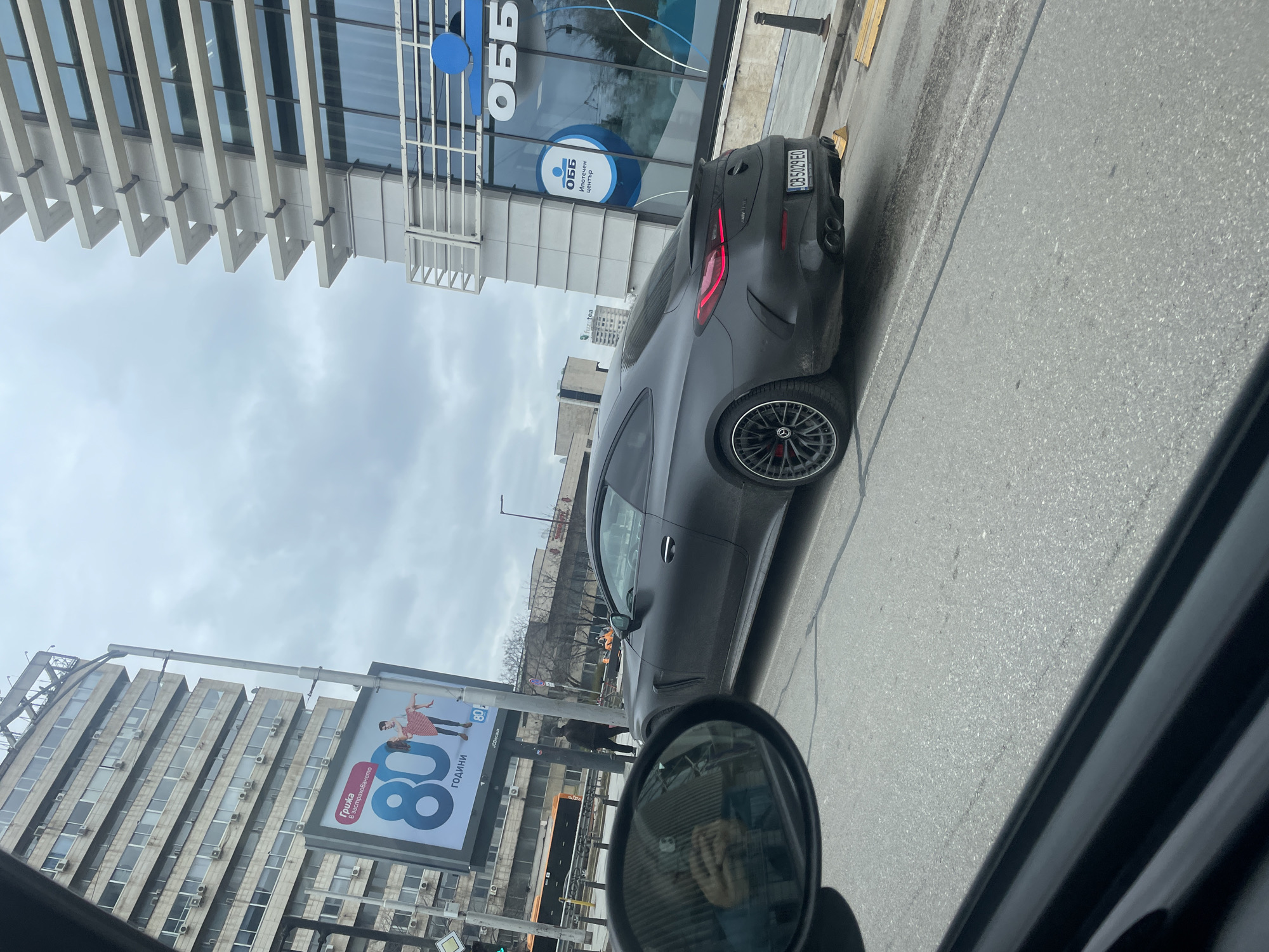 The image shows a street scene in Sofia, Bulgaria. A dark gray Mercedes-Benz AMG GT 4-Door Coupe is parked on the side of the road. The license plate is "CB 5029 EO". Behind the car is a modern building with the logo of "OBB" (United Bulgarian Bank). Across the street, an older building features an advertisement for an insurance company, celebrating "80 години" (80 years) and featuring the "JCDecaux" logo and an ad for "fuzetea." The image appears to have been taken from inside another car, as part of a side mirror and window frame are visible. The weather is overcast with a gray sky.