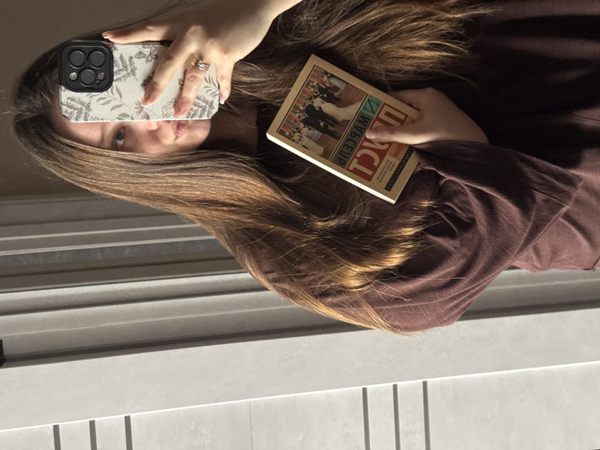 A female individual is depicted in an indoor setting located in Krasnodar, Russia, taking a mirror selfie. She has long, brown hair and is wearing a dark reddish-brown, long-sleeved top. Her right hand holds an iPhone with a light-colored botanical patterned case, featuring a three-lens camera module. Two rings are visible on the ring finger of her right hand, which has light-colored manicured fingernails. Her left eye is visible, and her lips are slightly puckered. Her left hand holds an open book with a light-colored cover featuring an illustration of multiple figures in formal attire. Mirrored text on the book cover includes "ПРИНЦ" (Prince). The background consists of a light-colored wall or paneling with horizontal dark lines.