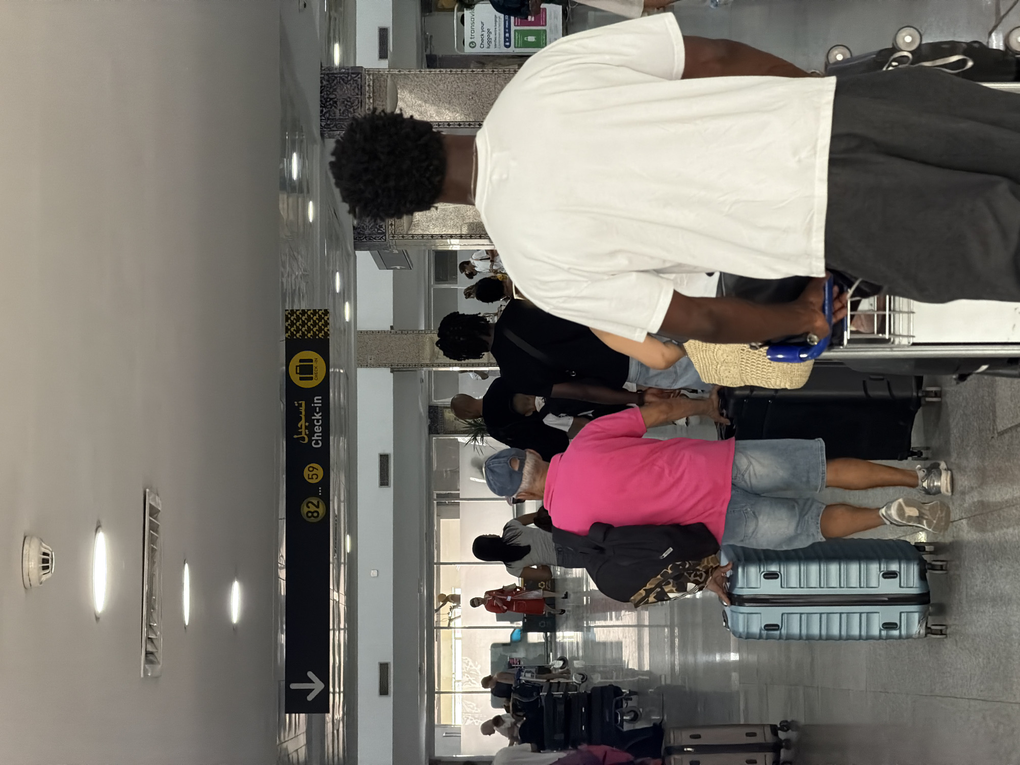 The image depicts the interior of an airport terminal in Marrakesh, Morocco, specifically within the departures or check-in area. Several individuals are visible, most of whom are walking with luggage. In the foreground, a Black man with dark curly hair is seen from behind, wearing a white t-shirt and dark pants, pushing a luggage cart. To his left, another man in a bright pink polo shirt and denim shorts is walking and pulling a light blue suitcase. Behind him, several other travelers with various suitcases and bags are moving through the concourse.
Overhead, a black sign with yellow text indicates "Check-in" with Arabic script above it, along with directional arrows and numbers "82" and "59," suggesting gate or counter numbers. The environment appears to be a modern airport terminal with tiled floors, recessed lighting, and large windows in the background, allowing natural light to enter. The overall atmosphere suggests a typical airport scene with passengers preparing for their flights.