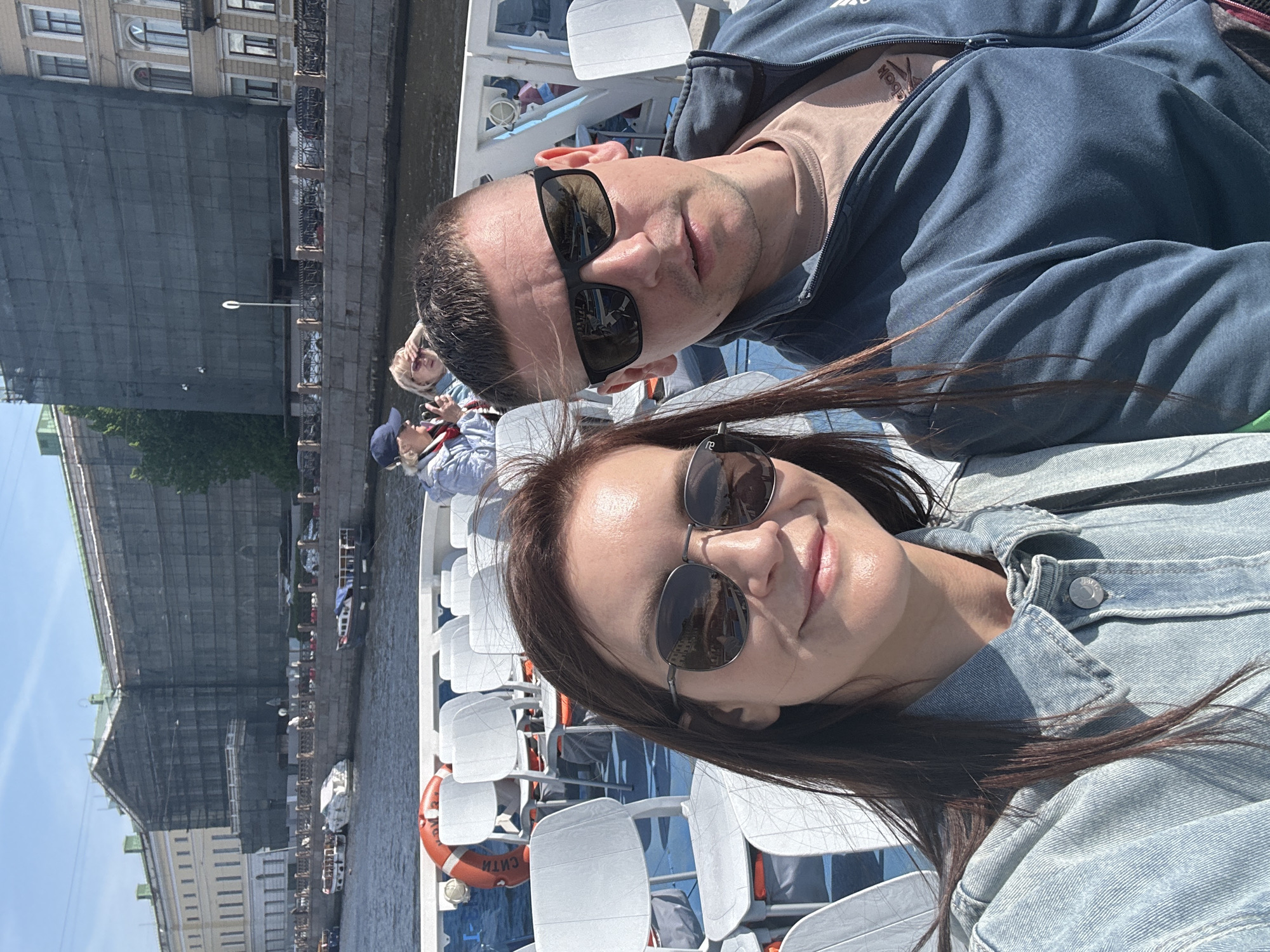 A medium outdoor shot shows a man and a woman in sunglasses on a boat with white chairs, apparently in Saint Petersburg, Russia. The man, up top, wears a blue jacket. The woman in the lower half wears a light blue jean jacket. The river is to the left, with the buildings lining the embankment in the background. A partial view of a life preserver is present on the left. Other people are in the background.