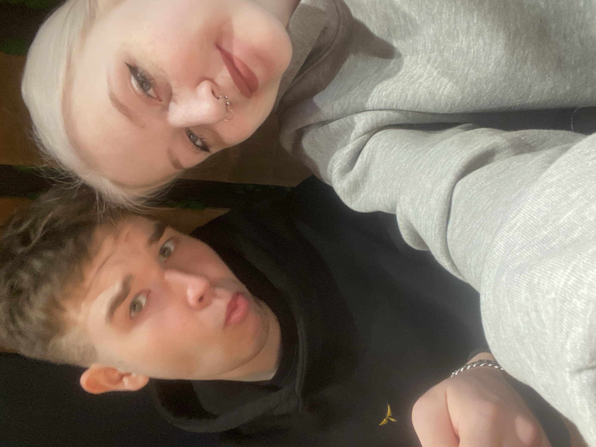 Two people are in the image, a woman and a man. The woman has light blonde hair and a nose ring. The man has short brown hair and is wearing a black hooded sweatshirt. The woman is wearing a gray hooded sweatshirt. The background is dark and indistinct. There is not enough information to determine the location as Izhevsk, Russia.