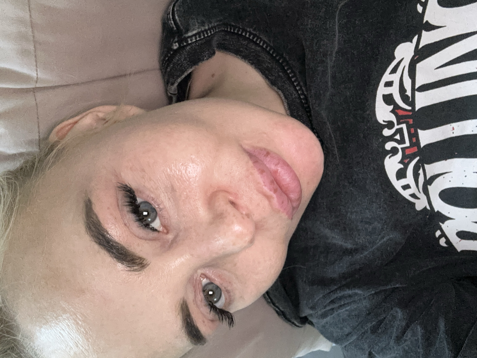 A close-up, angled portrait shows a woman with fair skin, light blonde hair, and gray eyes. She is wearing dark, possibly black clothing with white graphics visible on the right side of the image. Her eyebrows are prominent and appear to be cosmetically enhanced. The background appears to be a light pink fabric. The location is not discernible from the image. There is no indication of the city of Высокий, Russia.