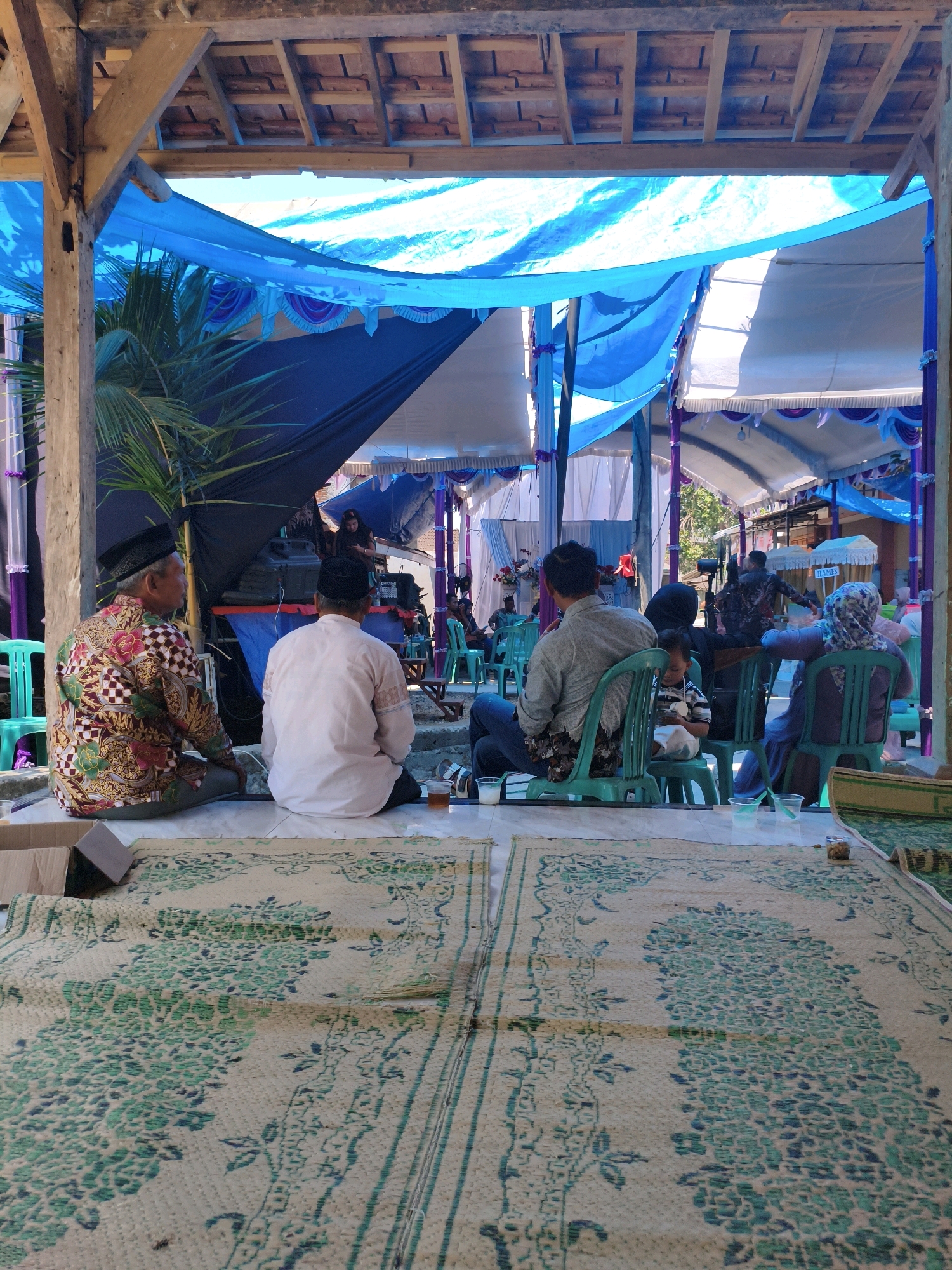 The image depicts an outdoor event, likely a gathering or ceremony, possibly in the city of Purwodadi, Indonesia. The scene is partially covered by a blue and white tarp structure supported by wooden beams, suggesting an open-air setting adapted for an event. In the foreground, two men are seated on woven mats on the ground, facing away from the camera. The man on the left wears a batik shirt with a colorful floral pattern and a black cap. The man on the right wears a plain light-colored shirt and a black cap. Between them are a few plastic cups and what appears to be a small pile of nuts.
Beyond the seated men, a group of people are visible sitting on green plastic chairs. Some are looking towards a stage or performance area in the background. This area has a dark backdrop and appears to have some equipment set up, possibly for music or presentation. Several other individuals are seen further back, some in shadow. The overall environment appears to be a community space or an outdoor venue decorated for a special occasion. The lighting suggests it is daytime.