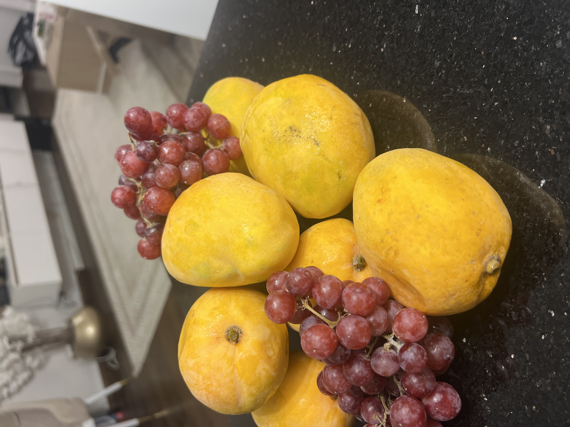 A still life of ripe mangoes and red grapes is displayed on a dark, speckled countertop. The fruit is artfully arranged in a cluster, with several mangoes forming the base and a bunch of grapes nestled amongst them. The mangoes are a vibrant yellow-orange, suggesting sweetness and ripeness. The grapes are a deep red, with a slight translucence that hints at their juicy interior. The background is softly blurred, showing elements of a modern interior, possibly in a residential setting in Sharm El Sheikh, Egypt, with hints of furniture and decor. The lighting is soft, highlighting the textures and colors of the fruit.