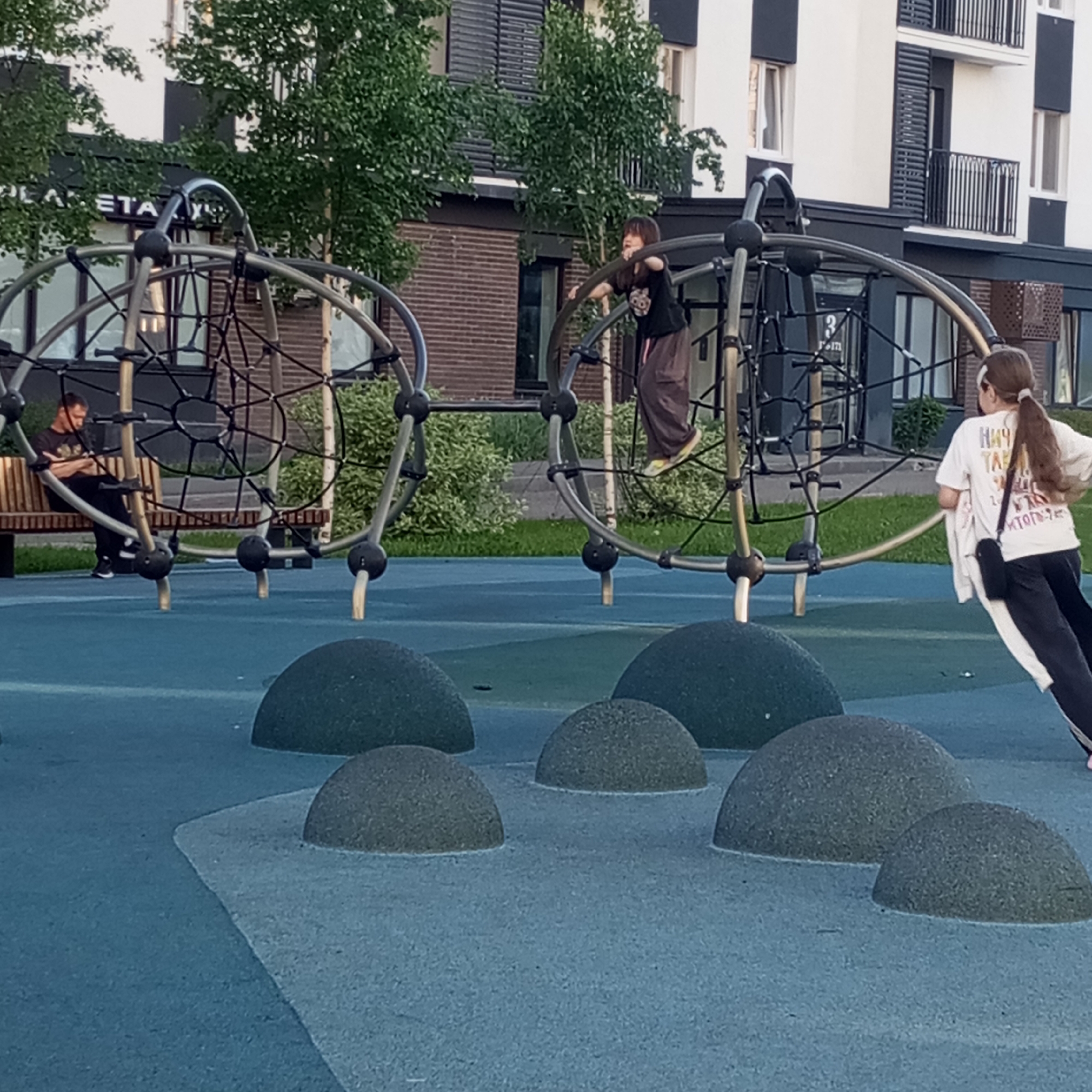 The image captures a late afternoon or early evening scene at a modern playground in Pervomaysky, Russia, with contemporary residential buildings in the background. The playground features a soft blue and grey ground, speckled with darker grey, half-dome-shaped elements.
In the foreground, two large, spherical climbing structures made of interconnected metal tubes and black rope nets dominate the scene. On the left, a man in a dark t-shirt and dark pants sits on a wooden bench beneath one of these structures, looking down, possibly at a phone. In the center, a child with long dark hair, wearing a dark t-shirt and brown pants with bright yellow shoes, is actively climbing the ropes of the rightmost structure, with both feet off the ground. On the far right, a young woman or girl with a long brown ponytail, dressed in a white t-shirt and dark pants, and carrying a black shoulder bag, stands facing away from the camera, seemingly observing the activities on the playground.
The background consists of multi-story residential buildings with distinctive white and dark grey/black facades, featuring numerous windows and balconies with vertical slat railings. Green trees are interspersed between the playground and the buildings. A sign partially visible on one of the buildings reads "PLANETA" (ПЛАНЕТА). On the white t-shirt of the person on the right, Cyrillic text is visible, including "НИЧУТА" and "КТОТО". The scene conveys a relaxed, casual atmosphere with individuals enjoying the outdoor space.